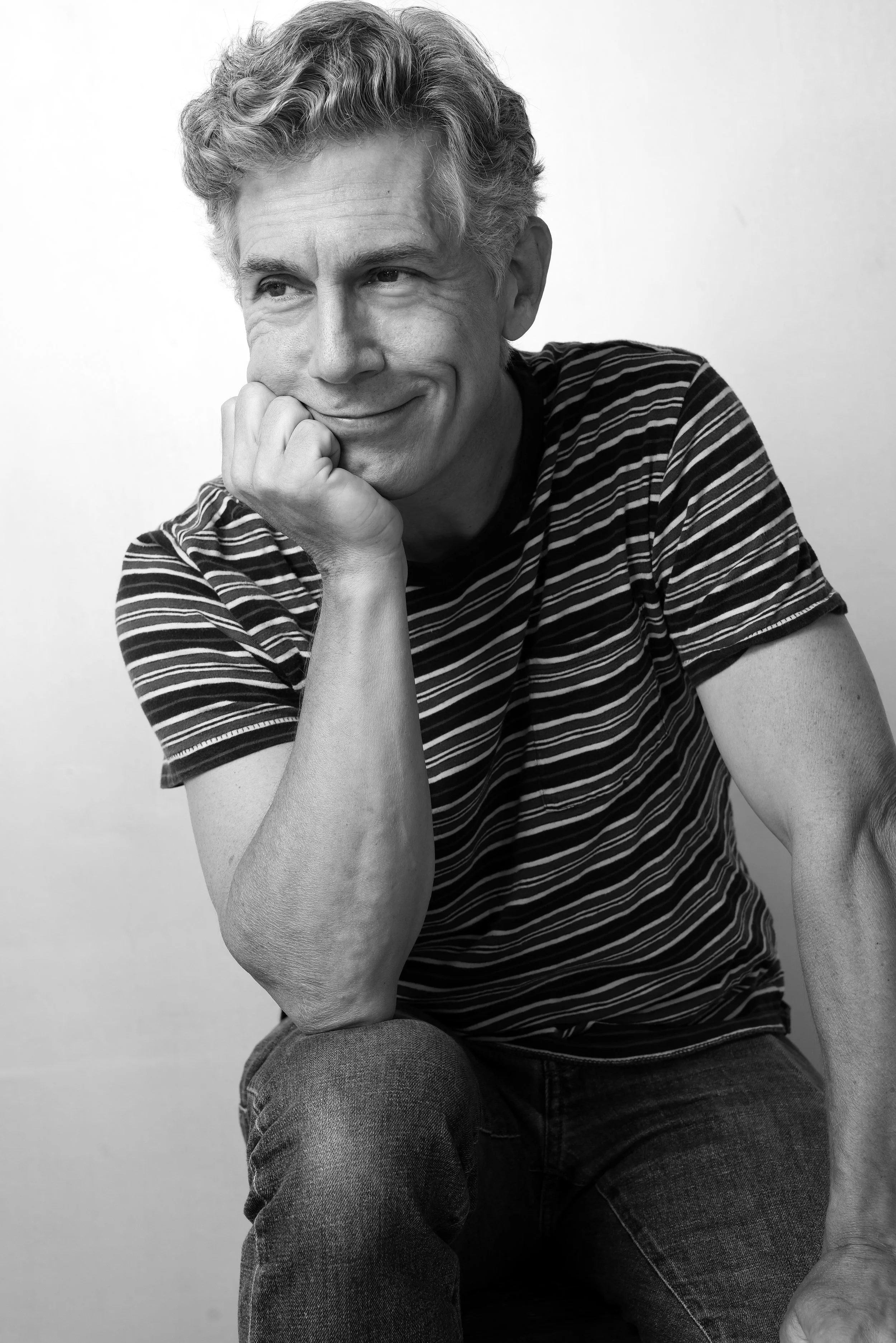 Black and white photo of a smiling man with curly hair, wearing a striped t-shirt, resting his chin on his hand.