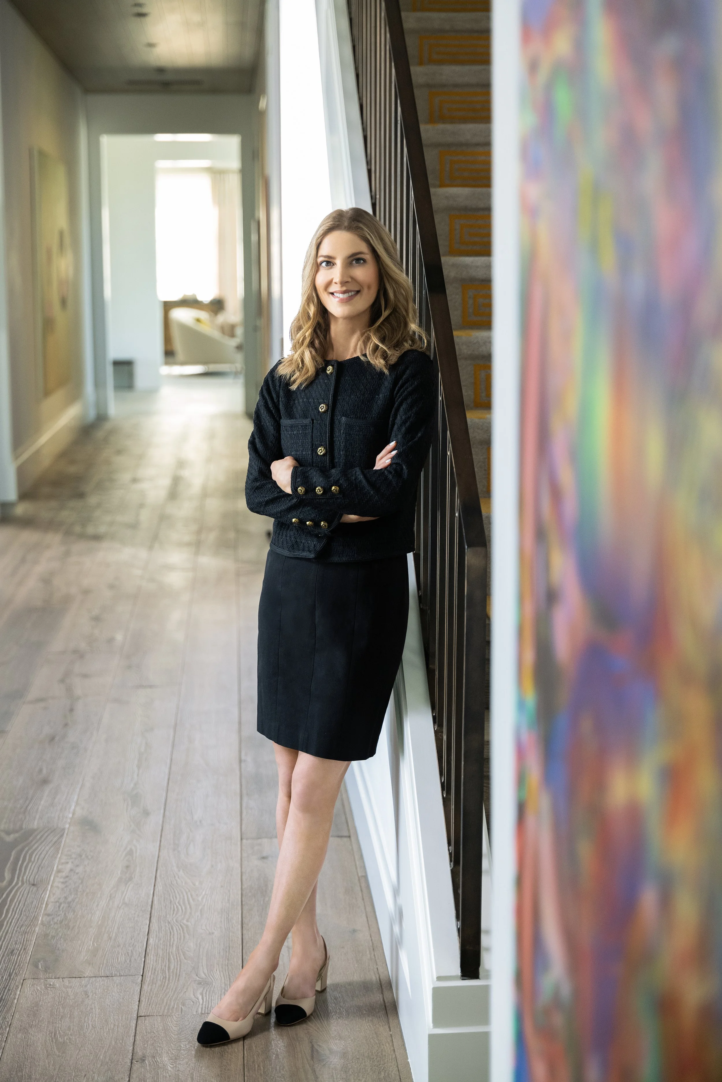 A woman with shoulder-length wavy blonde hair, wearing a black jacket with gold buttons and a black skirt, standing in a hallway with her arms crossed, smiling at the camera.