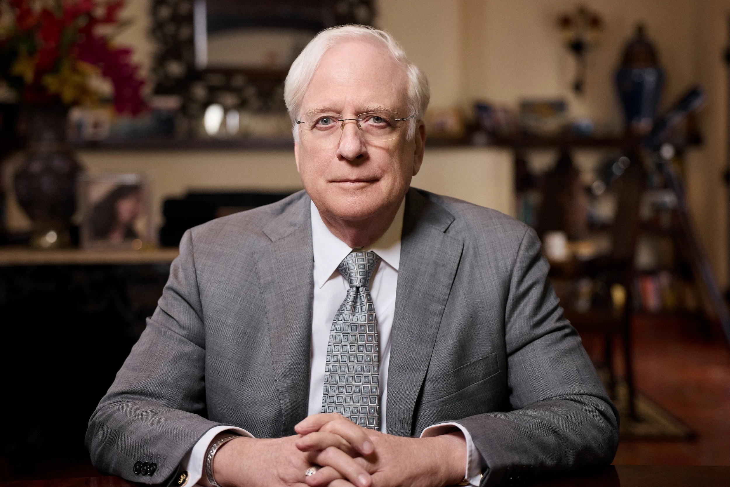 A middle-aged man with light-colored hair and glasses, wearing a gray suit and patterned tie, seated at a desk with his hands clasped, in a warmly lit room with shelves and decorative items in the background.  Image by top Los Angeles photographer Ke