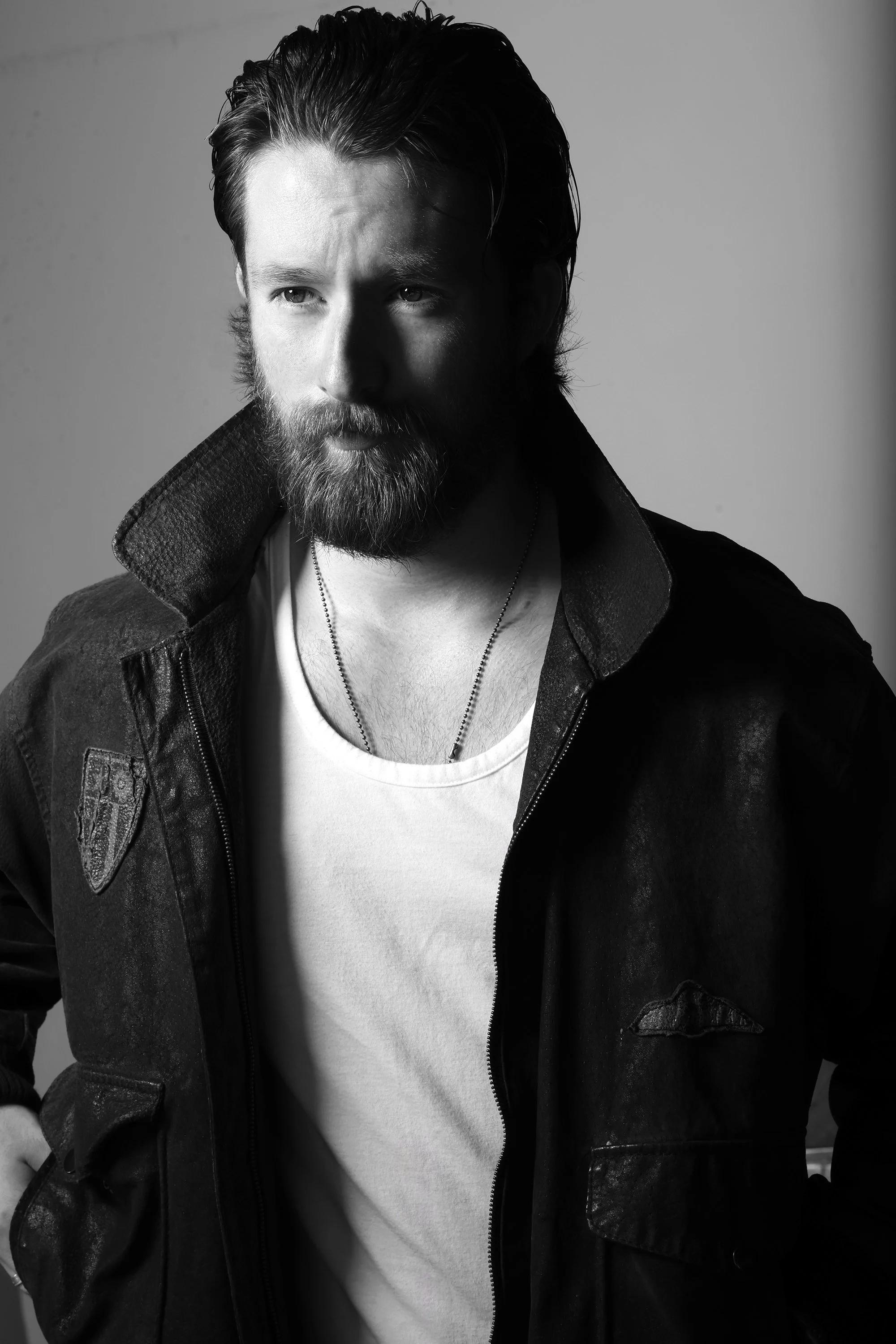 A black and white portrait of a man with a beard and styled hair, wearing a leather jacket over a white t-shirt and a chain necklace.   Image by top Los Angeles photographer Kenneth Dolin. 