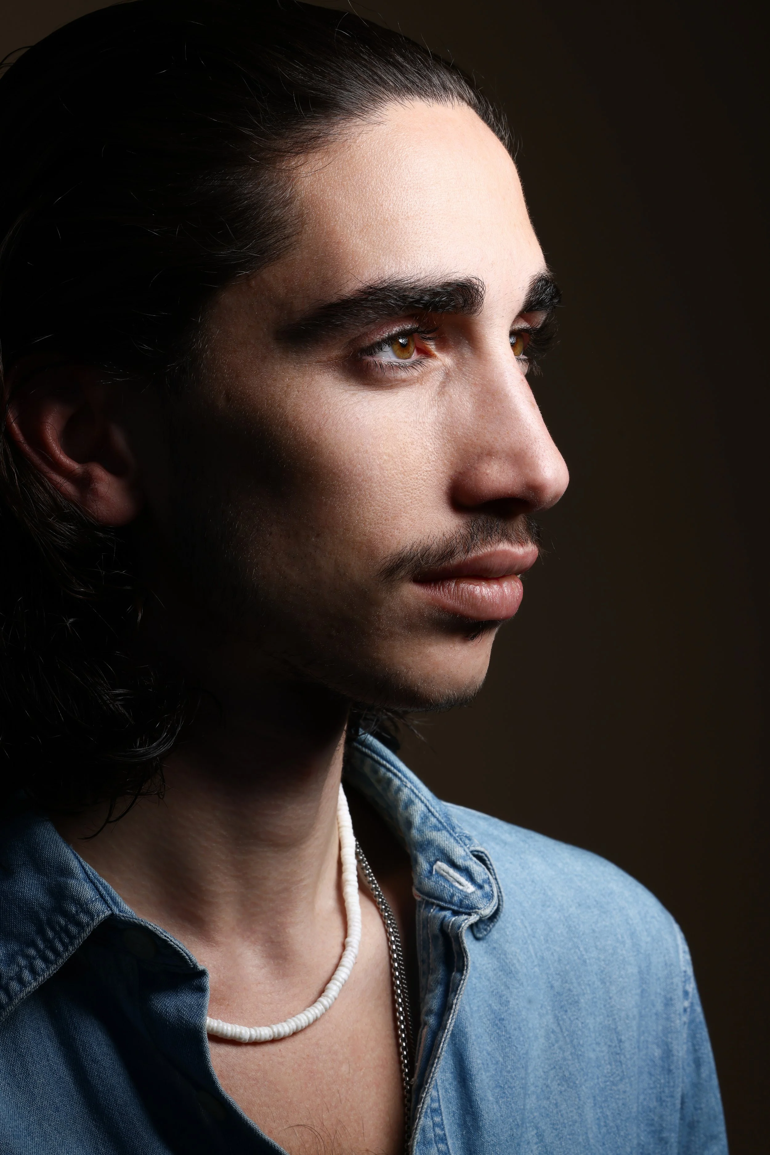 Close-up profile of a young man with long dark hair, light skin, brown eyes, wearing a blue denim shirt and a white beaded necklace, with a dark background.  Image by top Los Angeles photographer Kenneth Dolin. 