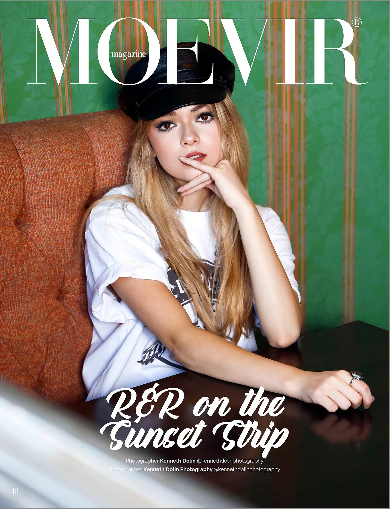 A young woman with long blonde hair, wearing a black beret and a white t-shirt, sitting at a table with a colorful background, looking at the camera with her finger on her lips.