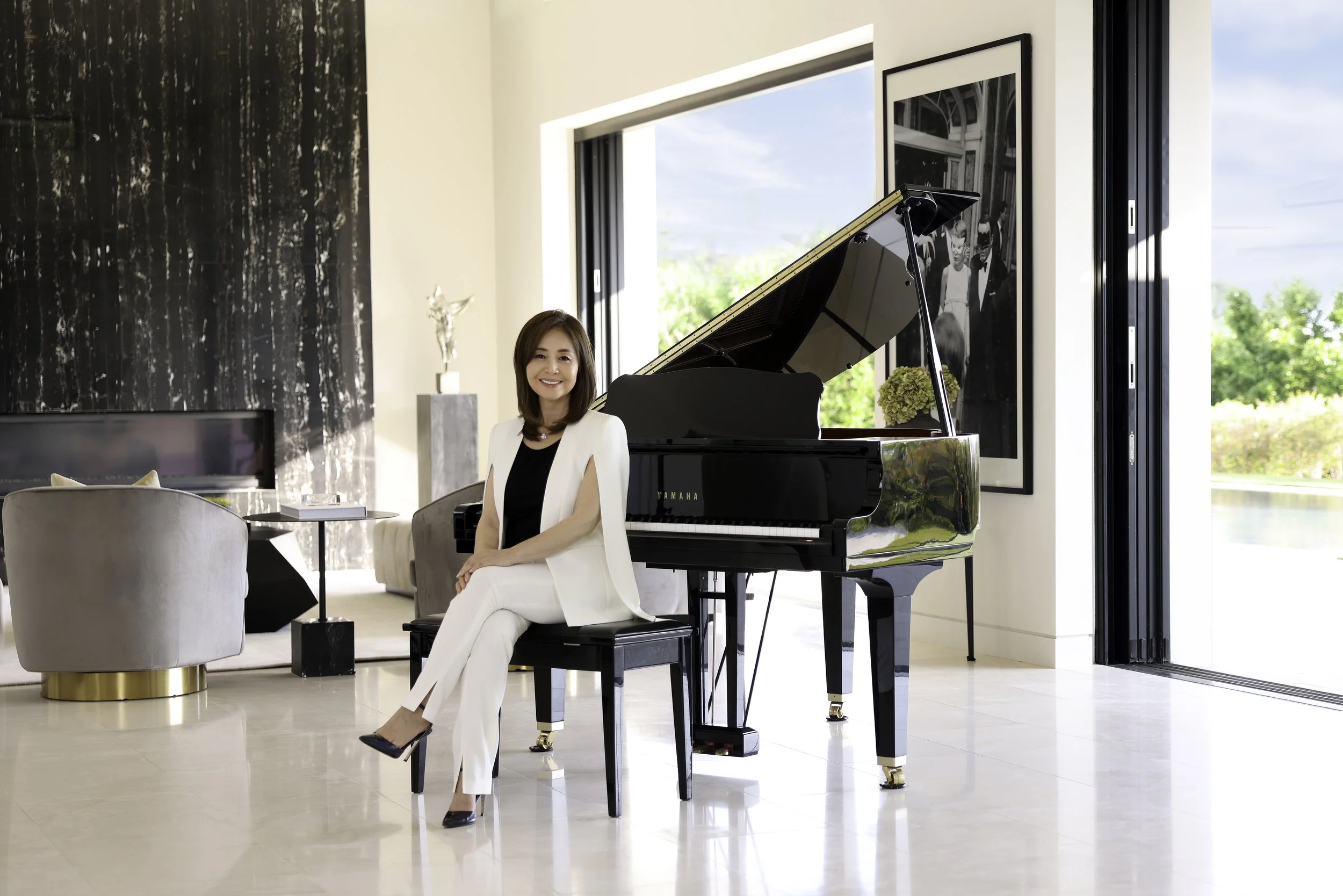 A woman in white pants and a white blazer sitting on a piano bench next to a black grand piano in a modern living room.