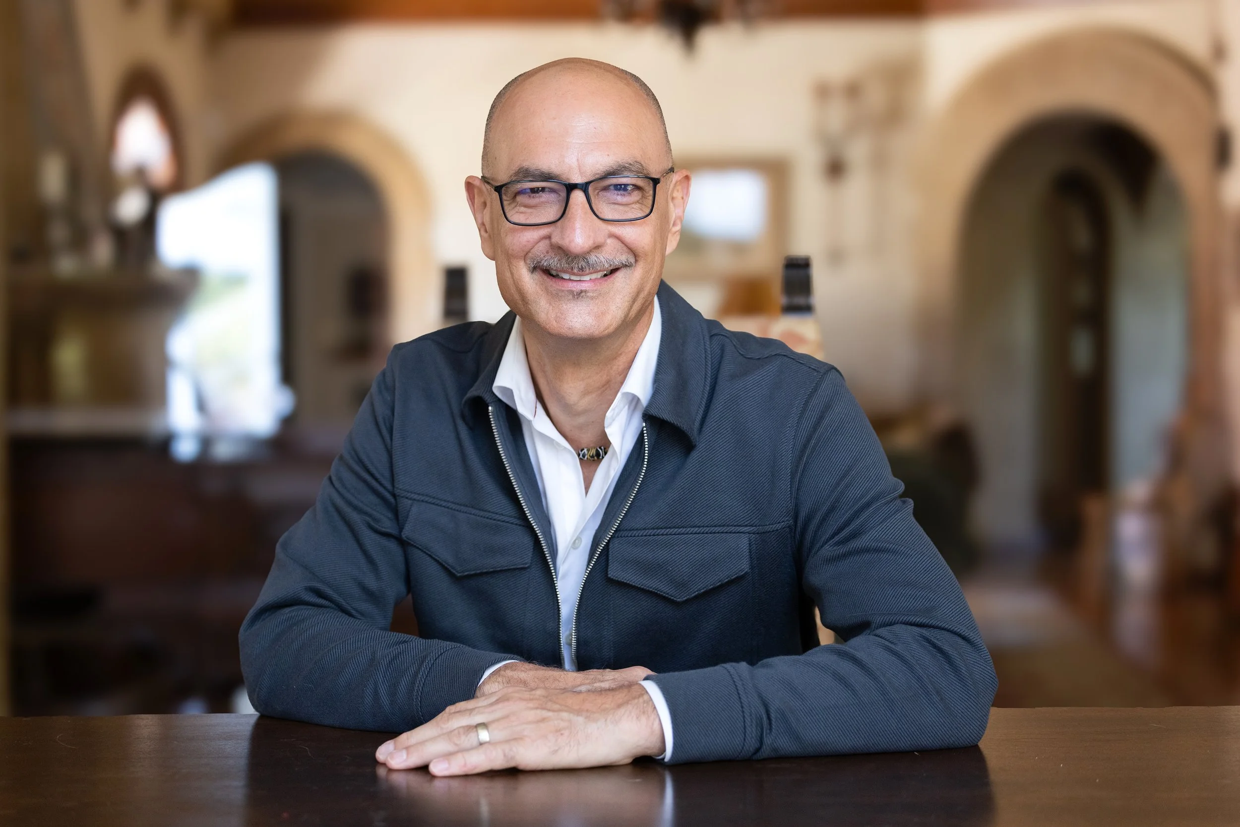 An older man with glasses, a smile, and a bald head, wearing a white shirt and a black jacket, sitting at a wooden table in a well-lit, cozy room with arched doorways.  Image by top Los Angeles photographer Kenneth Dolin. 