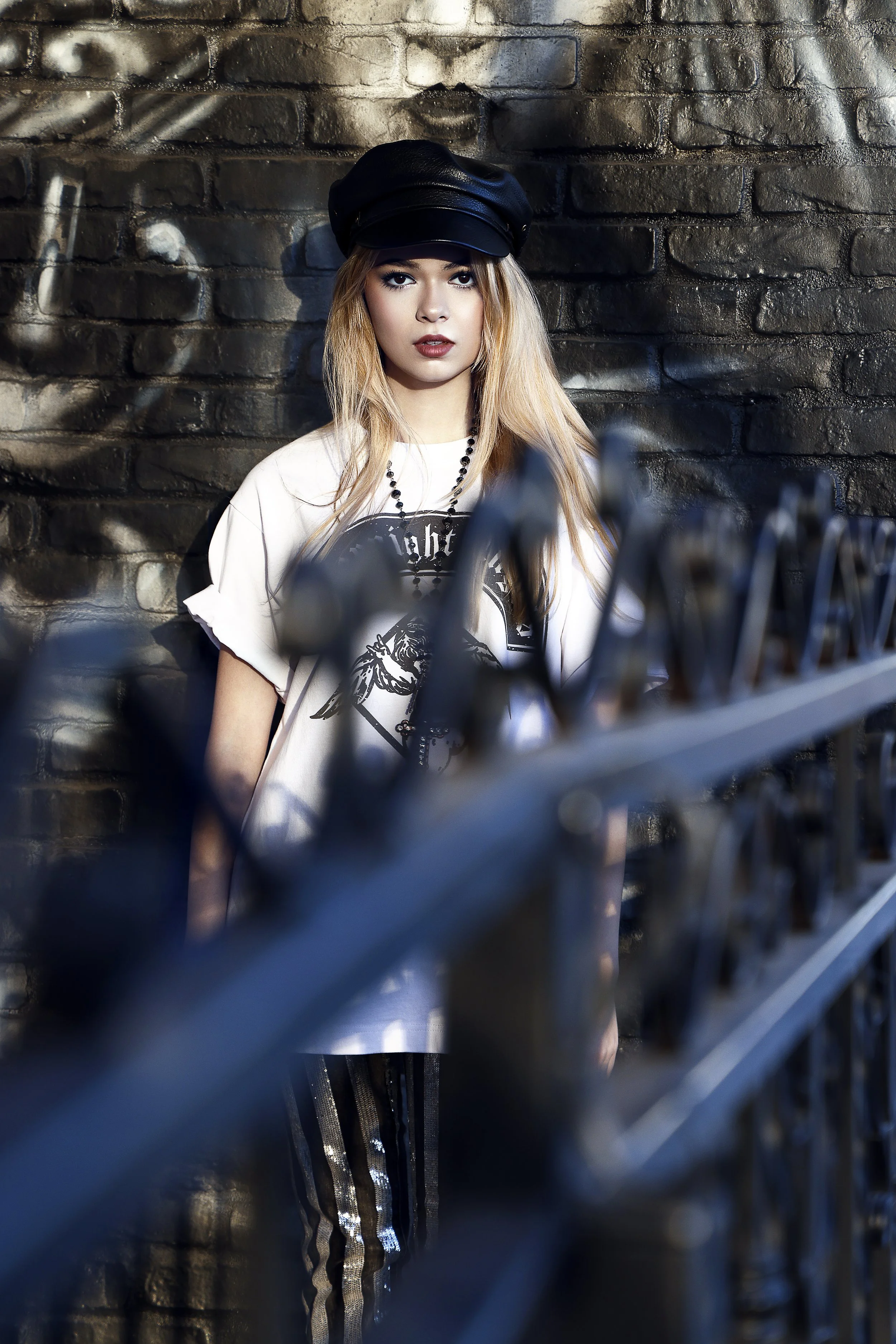 A young woman with long blonde hair wearing a black cap, a white graphic t-shirt, black bead necklace, and black and silver striped pants, standing against a dark brick wall.