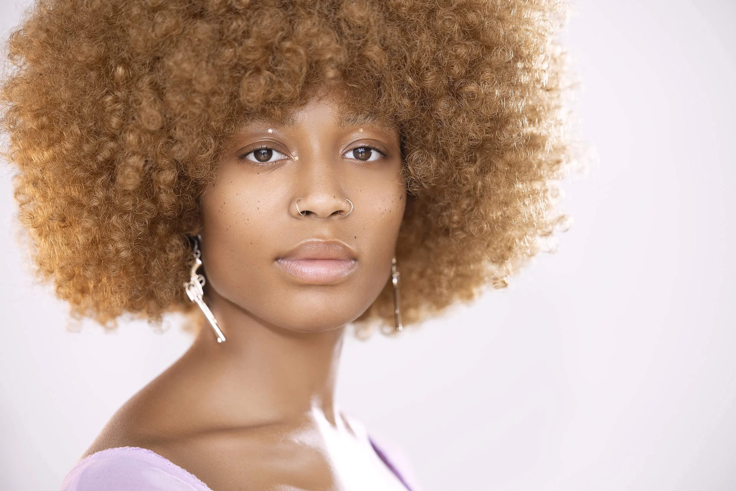 Close-up of a young woman with a large, curly, blonde afro hairstyle. She has light skin with freckles, wearing subtle makeup, a nose ring, and drop earrings. She is looking directly at the camera with a neutral expression.