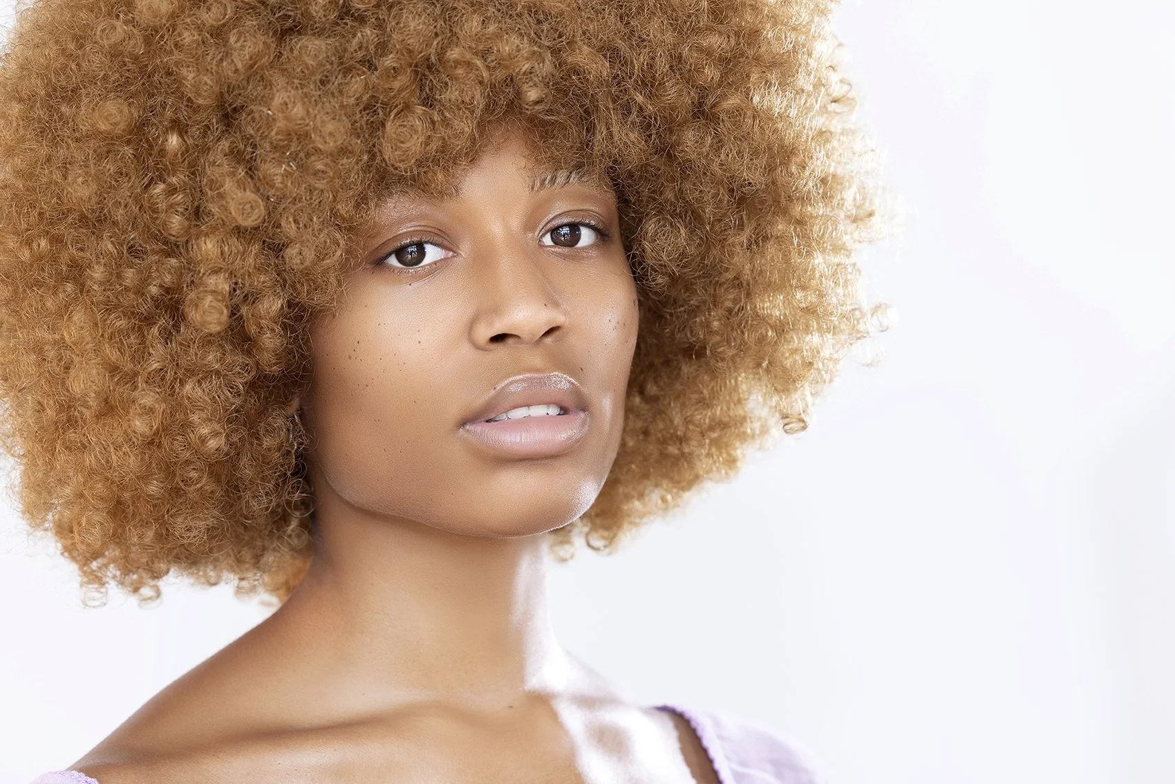 Close-up of a young woman with a large, curly, blonde afro hairstyle. She has light skin with freckles, wearing subtle makeup, a nose ring, and drop earrings. She is looking directly at the camera with a neutral expression.