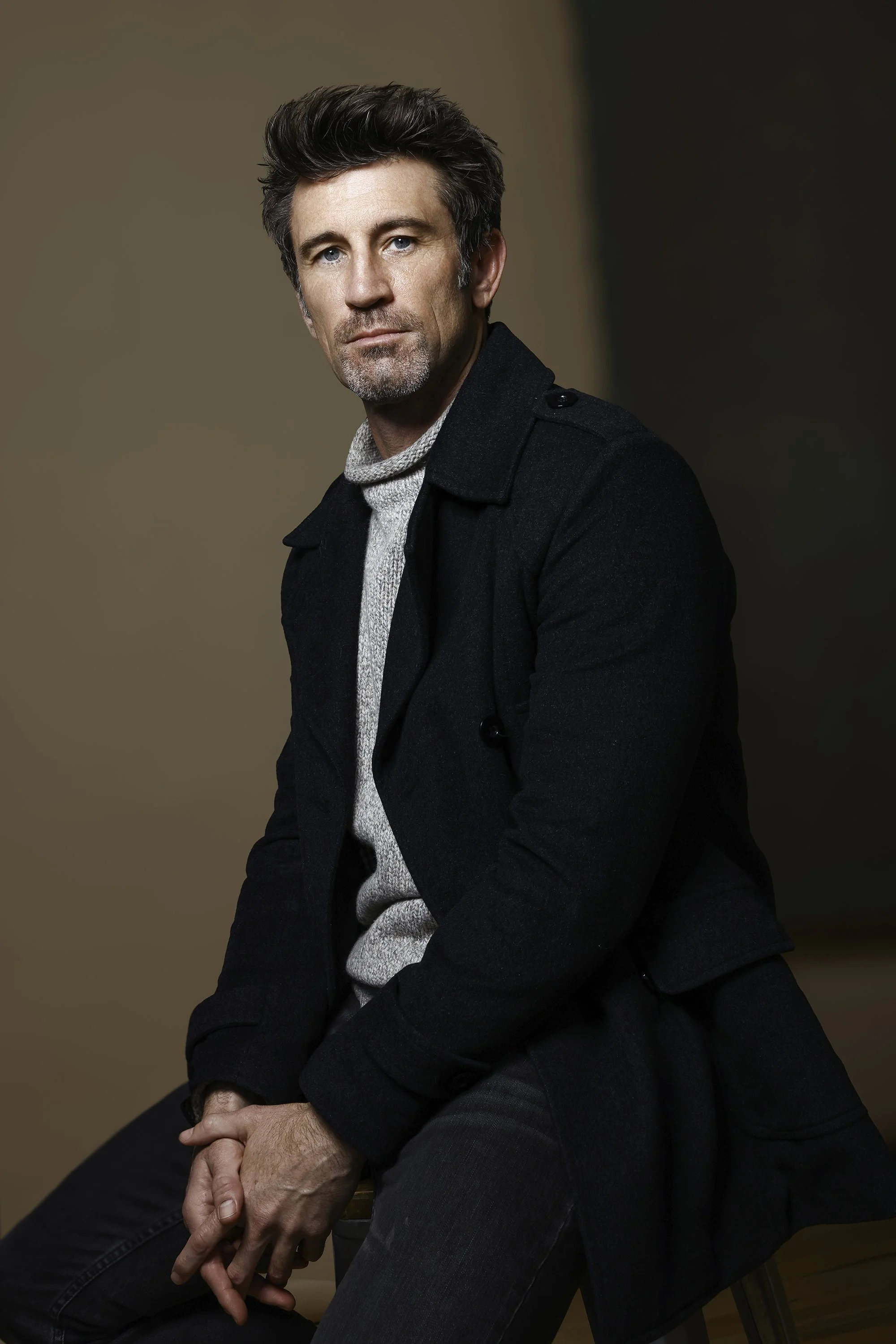 A middle-aged man with dark hair and a beard, wearing a dark coat over a light knit sweater, sitting on a chair against a plain brown background.  Image by top Los Angeles photographer Kenneth Dolin. 