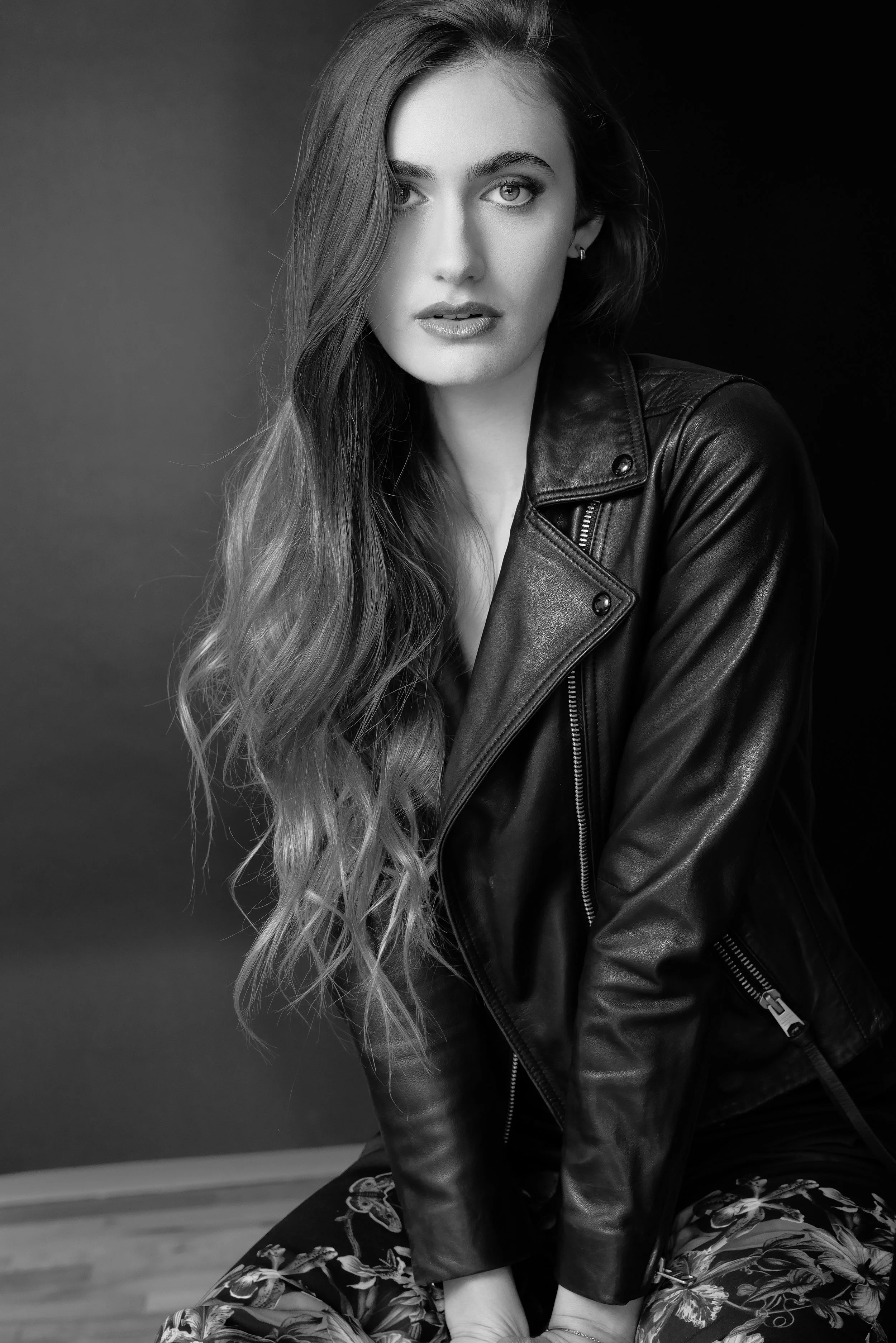 Black and white portrait of a woman with long, wavy hair wearing a leather jacket, sitting on the floor against a dark background.   Image by top Los Angeles photographer Kenneth Dolin. 