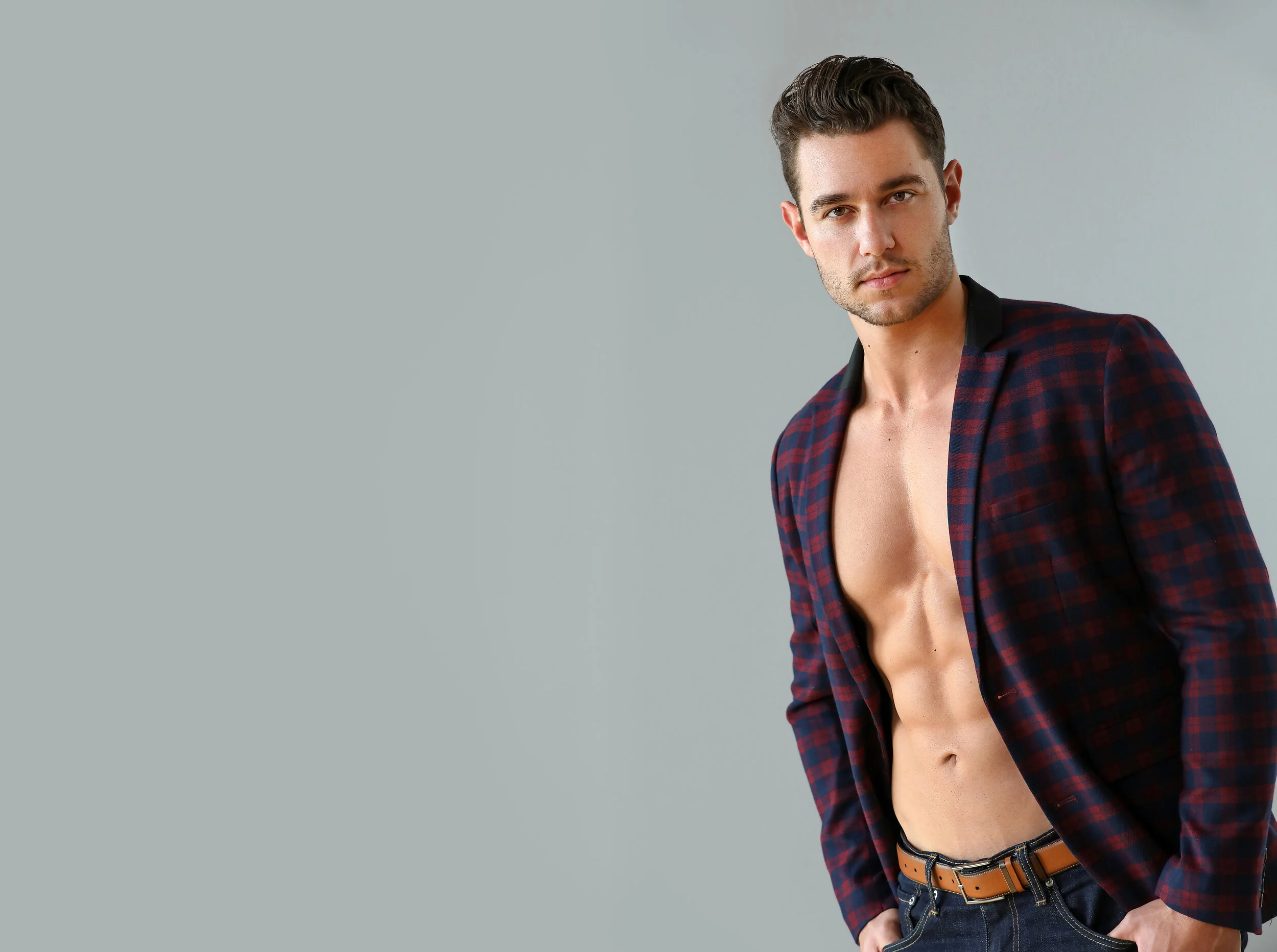 A young man with dark hair and light skin standing against a light gray background, wearing an unbuttoned dark red and navy plaid shirt revealing his bare chest and abs, with his left hand in his pocket and looking at the camera.