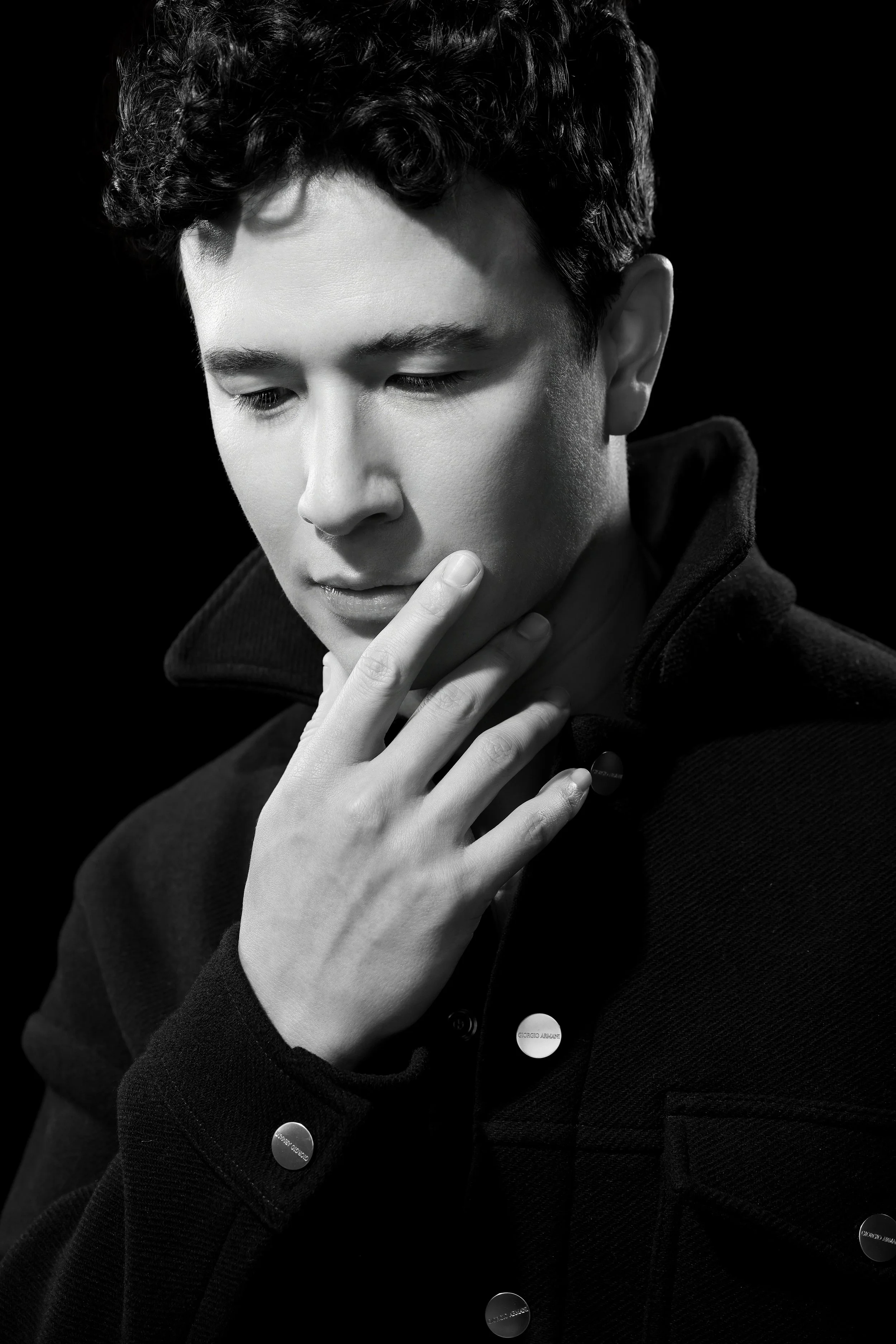 Black and white portrait of a young man with curly hair, looking downward with his hand touching his chin, wearing a jacket with visible buttons.  Image by top Los Angeles photographer Kenneth Dolin. 