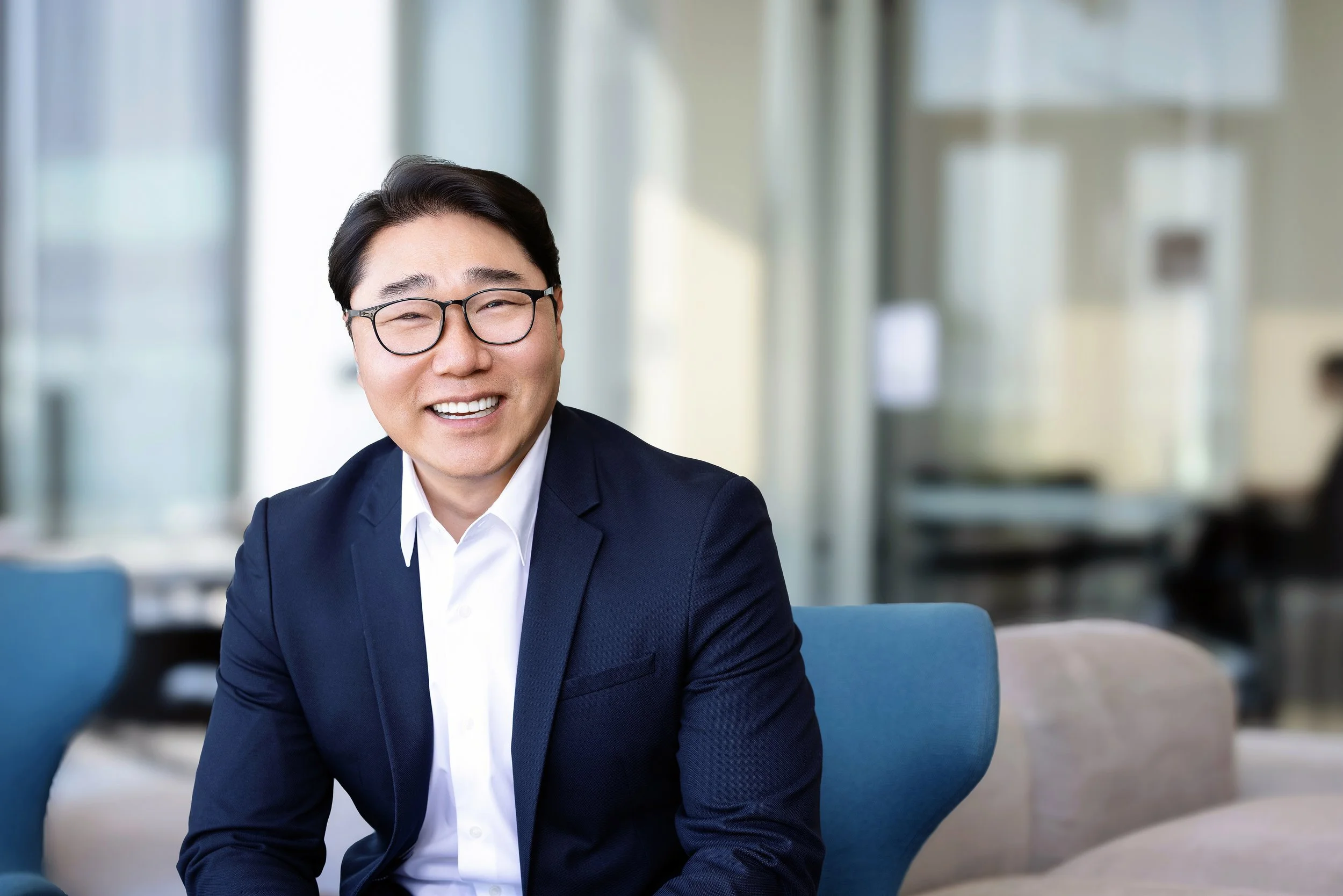 A smiling man in a suit and glasses sitting on a blue chair in an office setting.