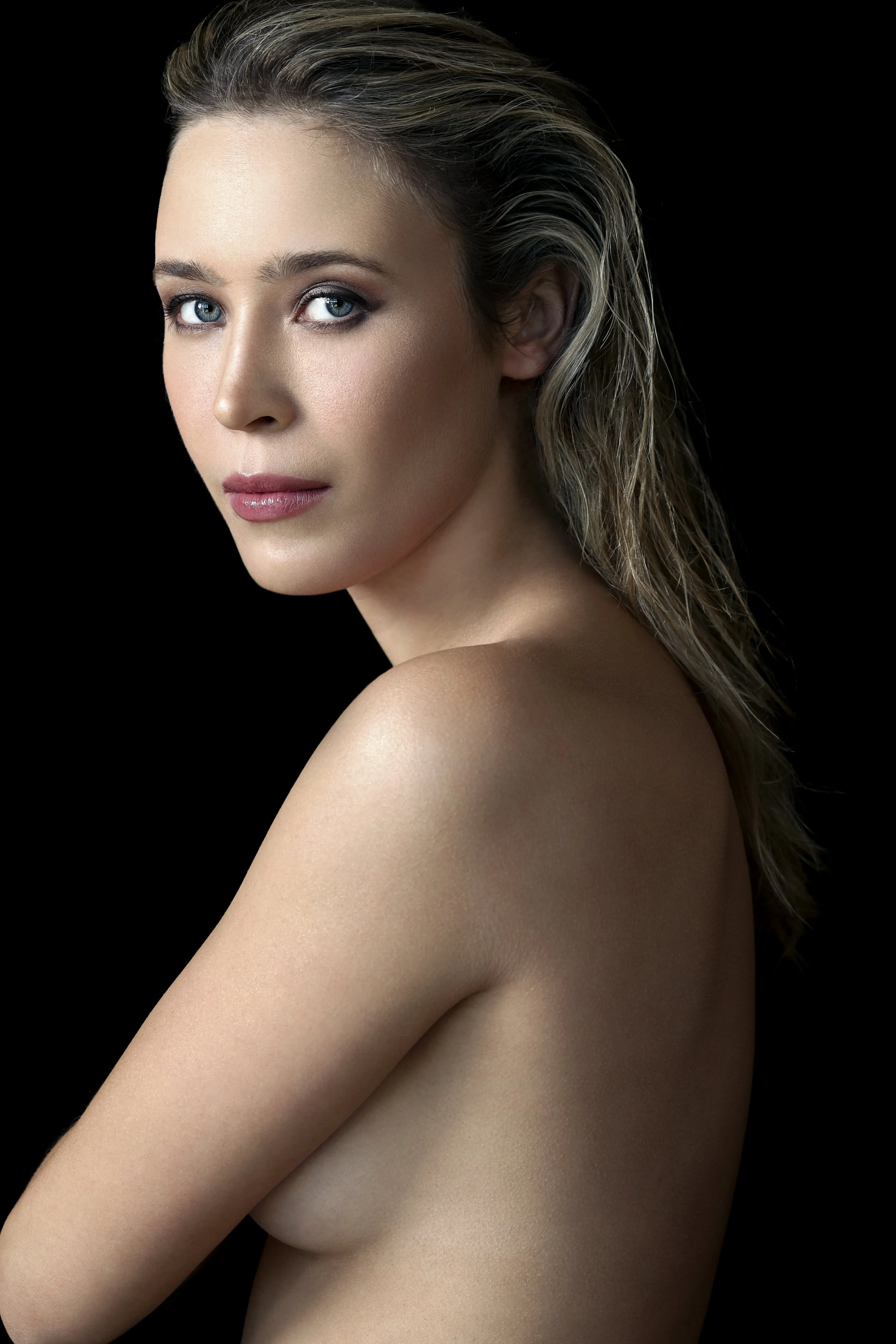 A portrait of a woman with blue eyes, light makeup, and wavy blonde hair, looking at the camera against a black background.