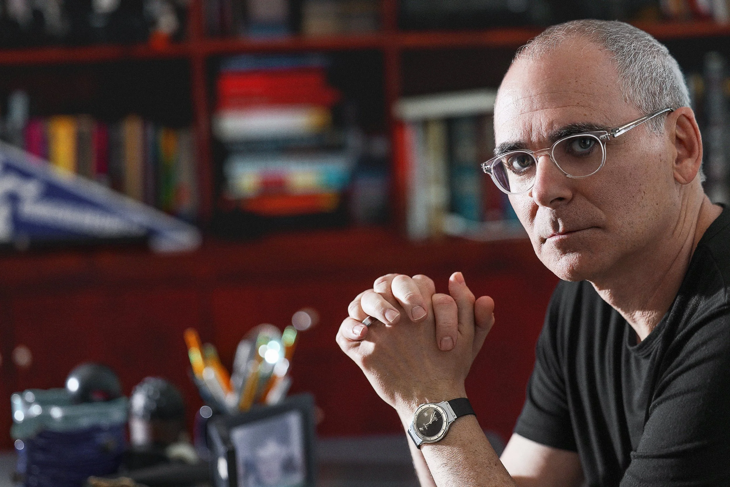 A man with glasses and a watch sitting at a desk with hands clasped, looking at the camera in a room with shelves of books and objects in the background.  Image by top Los Angeles photographer Kenneth Dolin. 