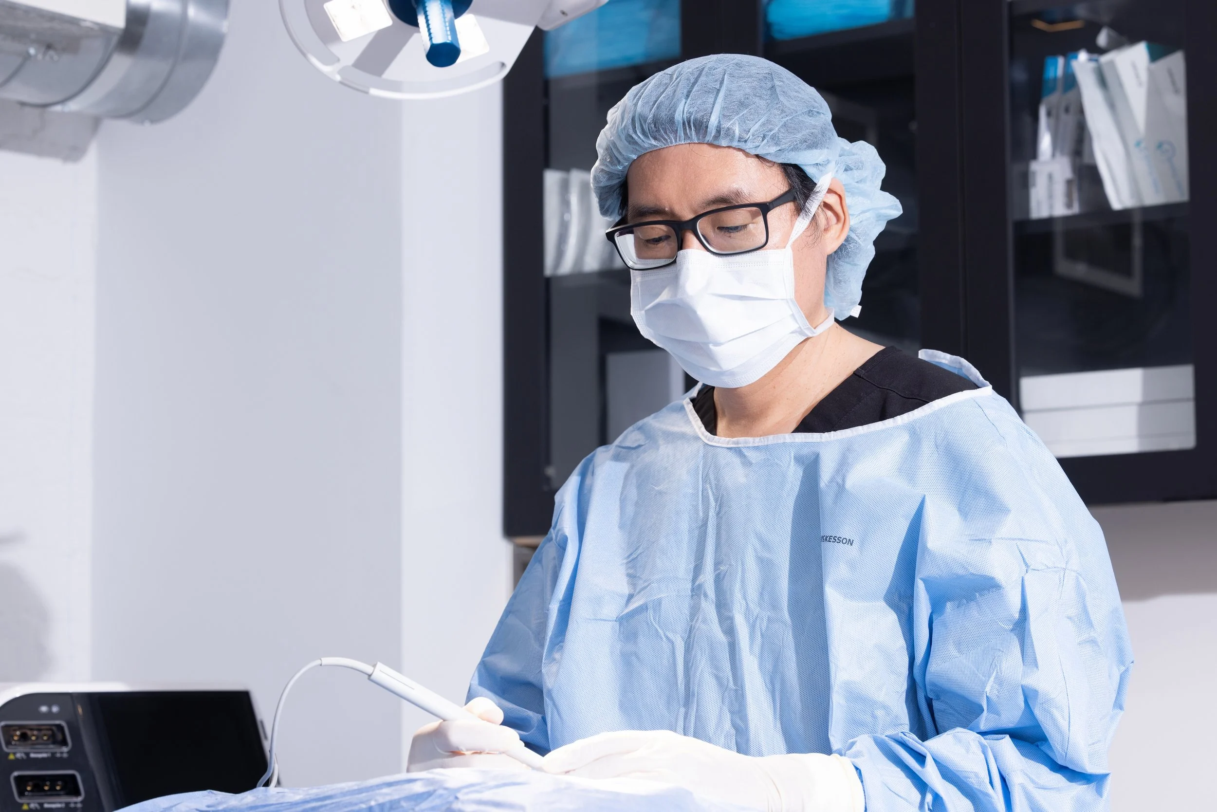 A medical professional in scrubs, a surgical mask, a hair cap, and glasses performing a procedure in a clinical setting.
