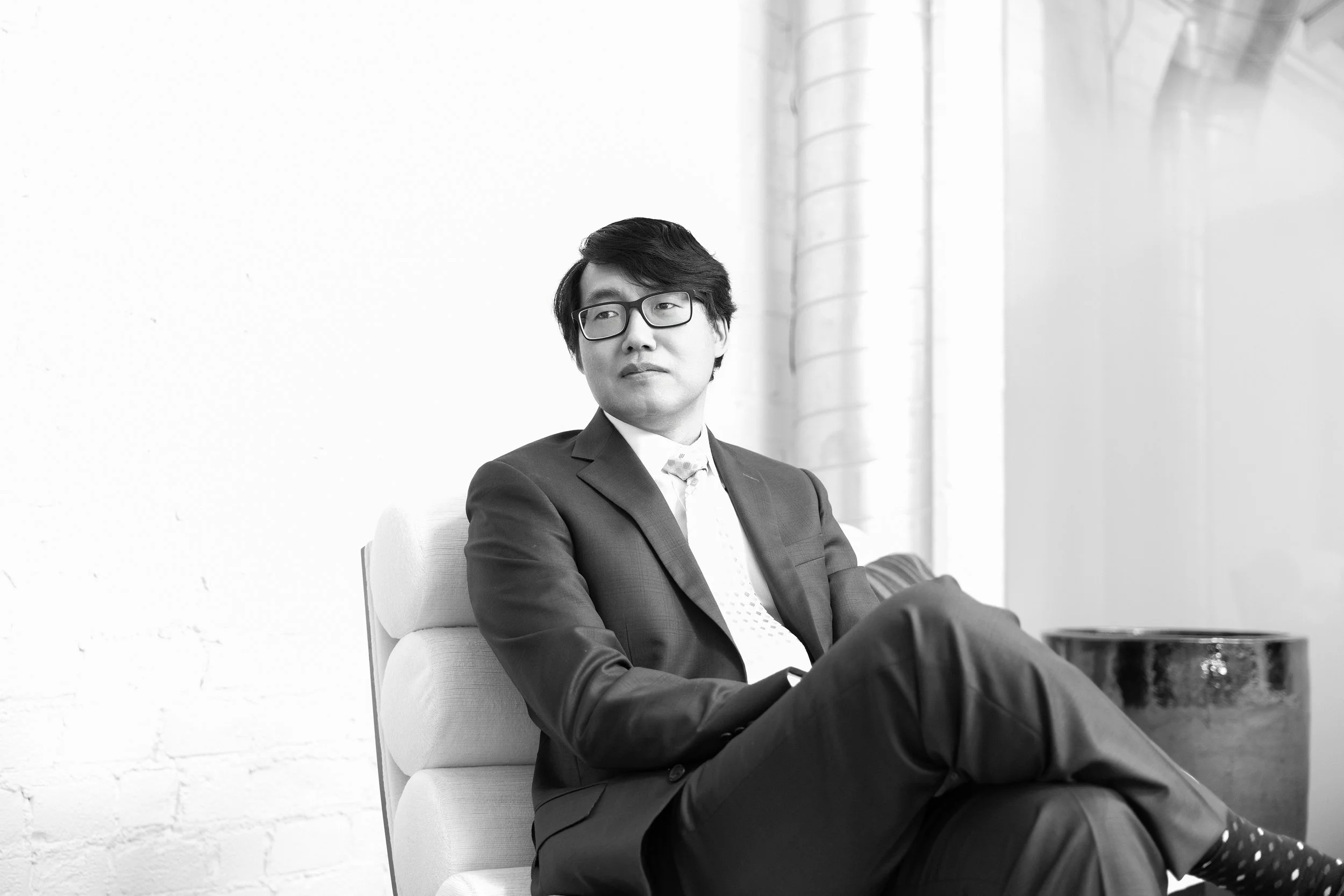 A young man in a suit and glasses sitting on a cushioned chair, looking to the side, in a well-lit room with a white wall and window with curtains.