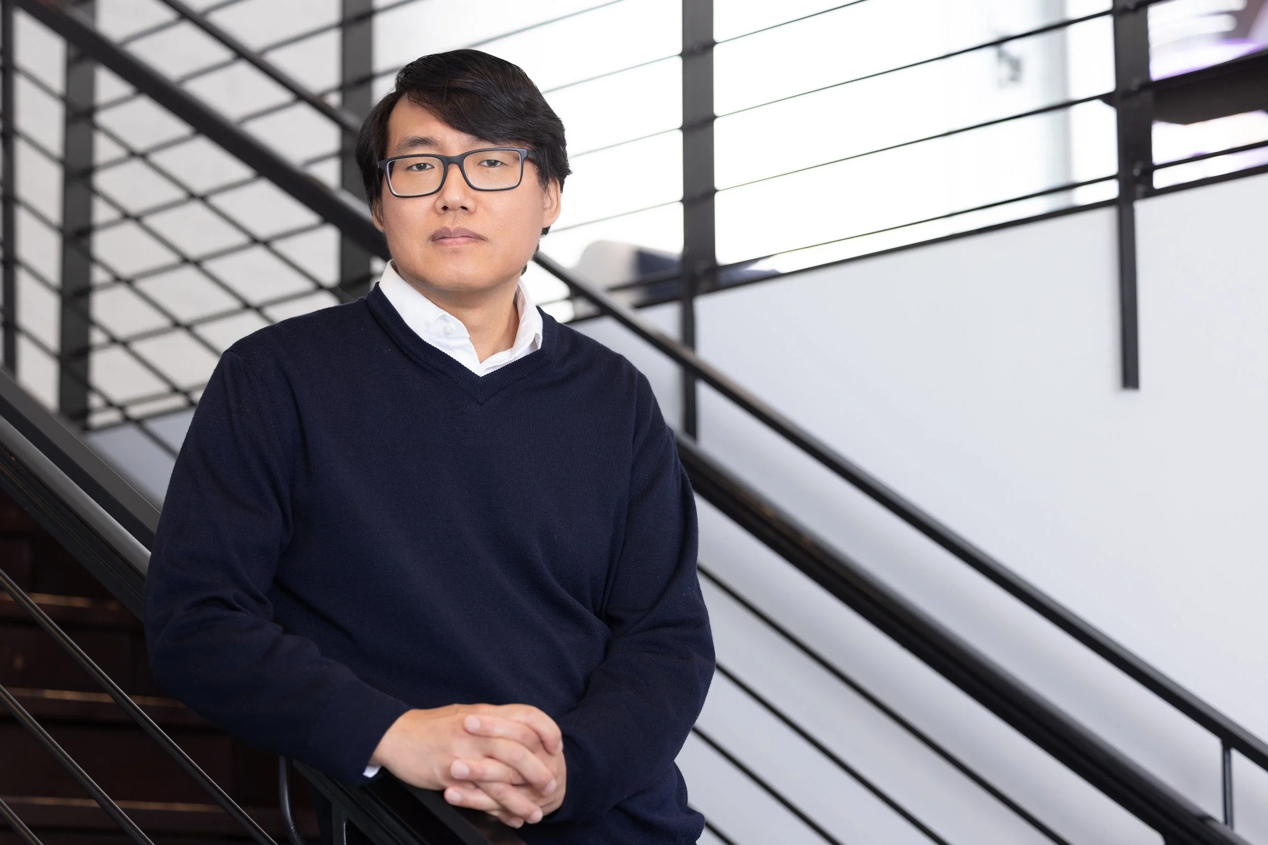 A young man with black hair, wearing glasses, a white collared shirt, and a navy blue sweater, standing on a staircase with black metal railings and large windows in the background.