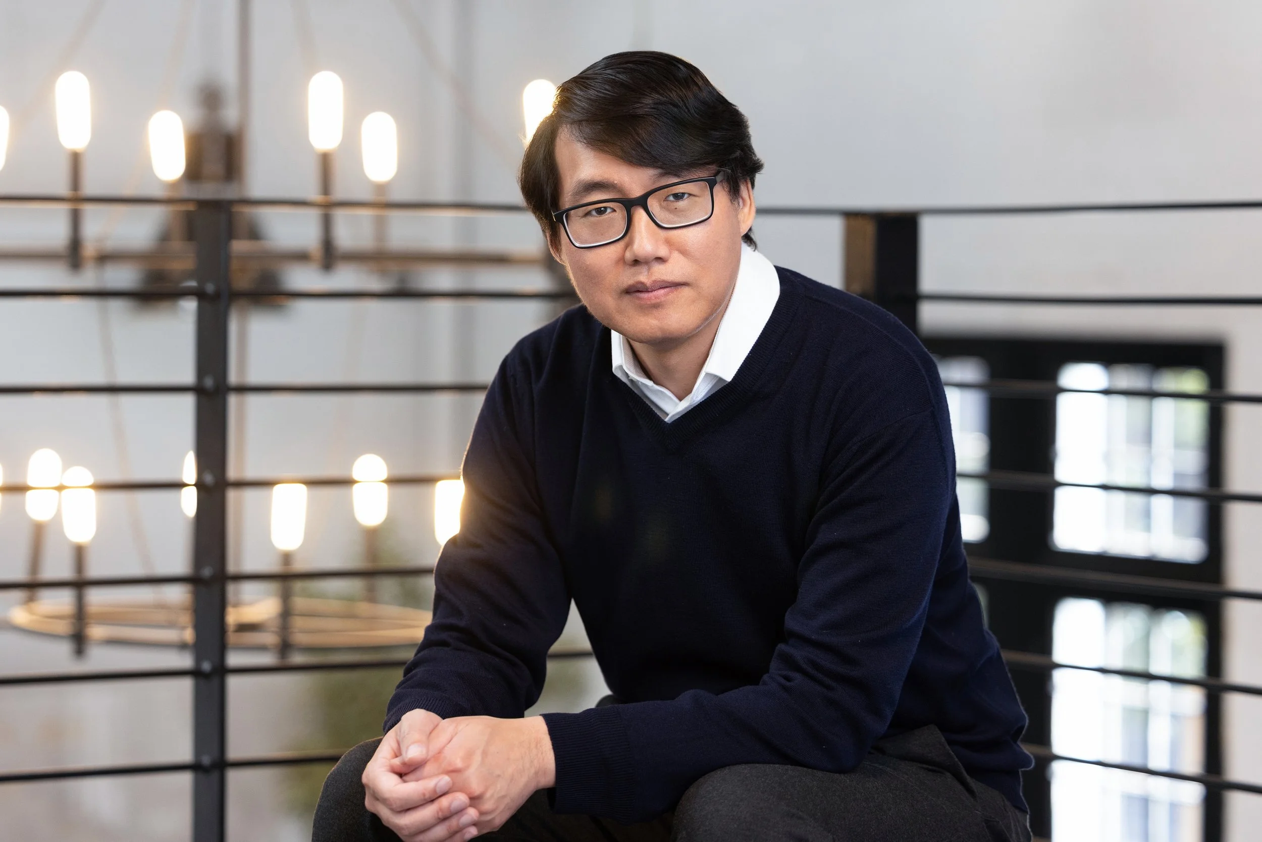 A man with black hair, glasses, wearing a white shirt and navy sweater, sitting indoors near a black metal railing and lit bulbs, looking at the camera.