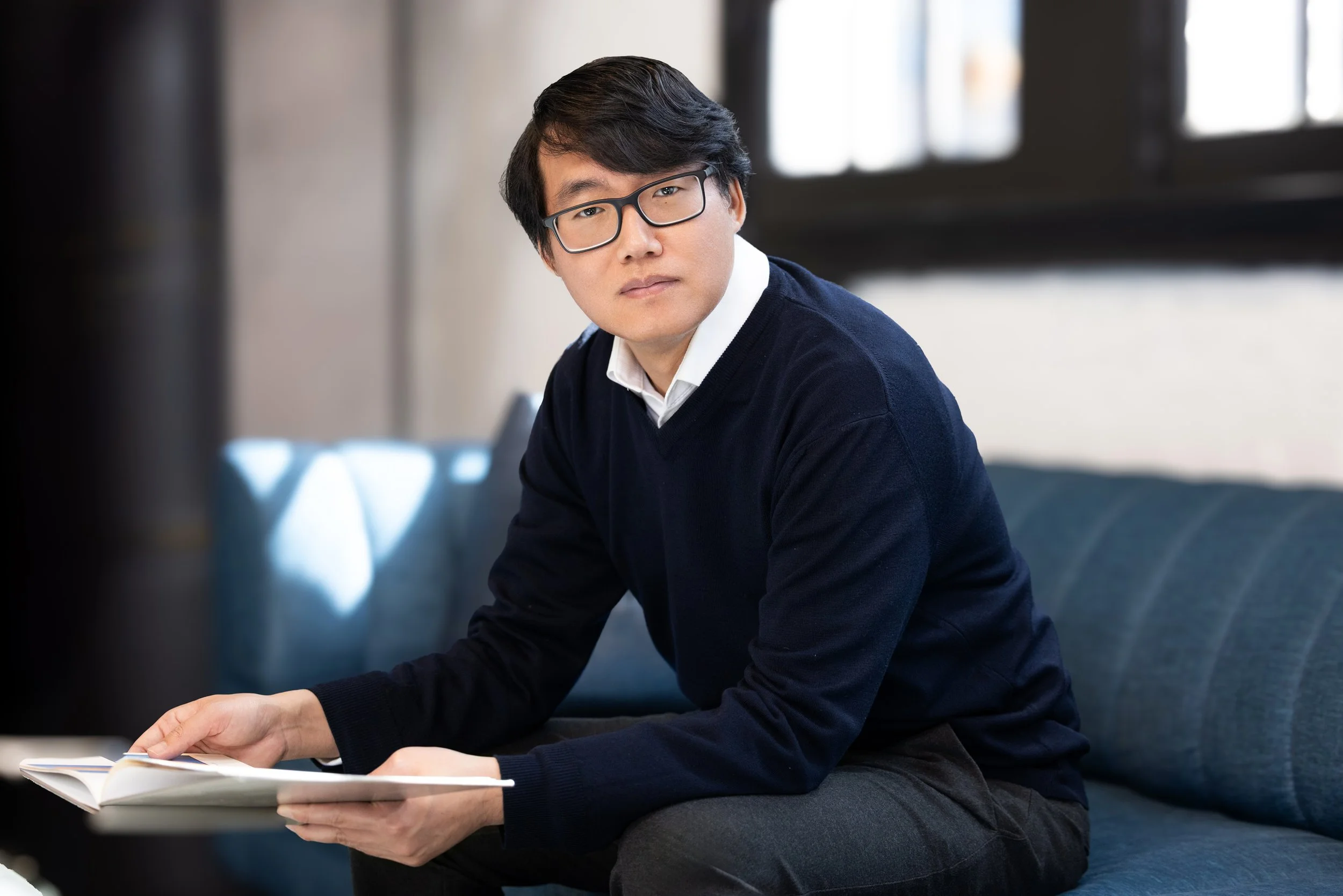 A doctor in his office sits on a blue couch holding an open book, looking towards the camera with a serious expression. Photo by Kenneth Dolin Photography, the top photographer in Los Angeles. 