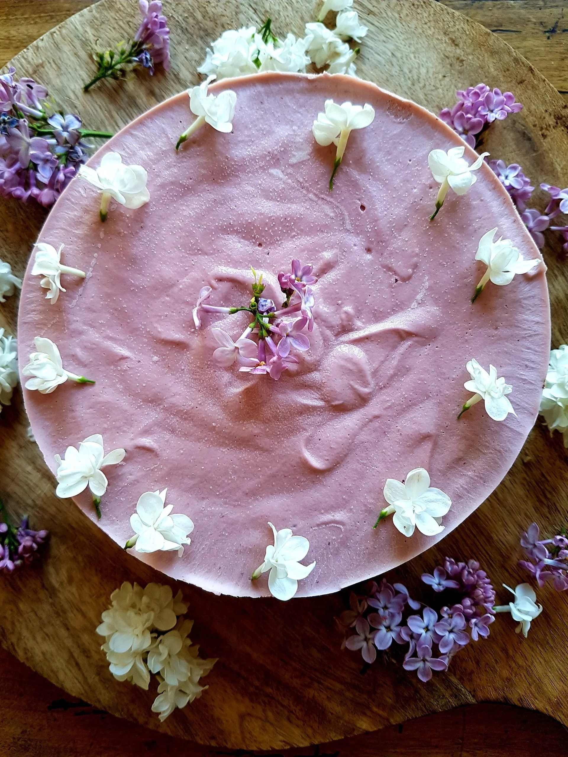 Pink raw cake decorated with white and purple lilac flowers on a wooden board