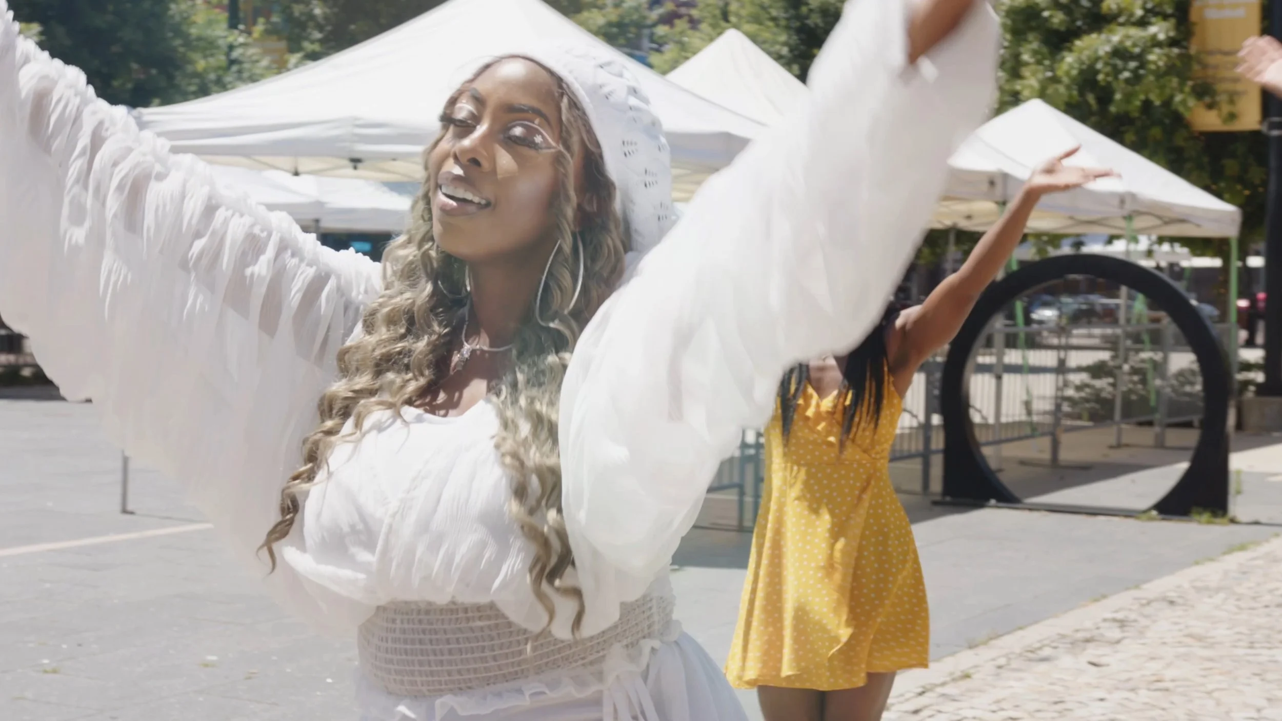 Two women dancing outdoors at a festival, one in a white dress and headscarf, the other in a yellow polka dot dress, with tents in the background.