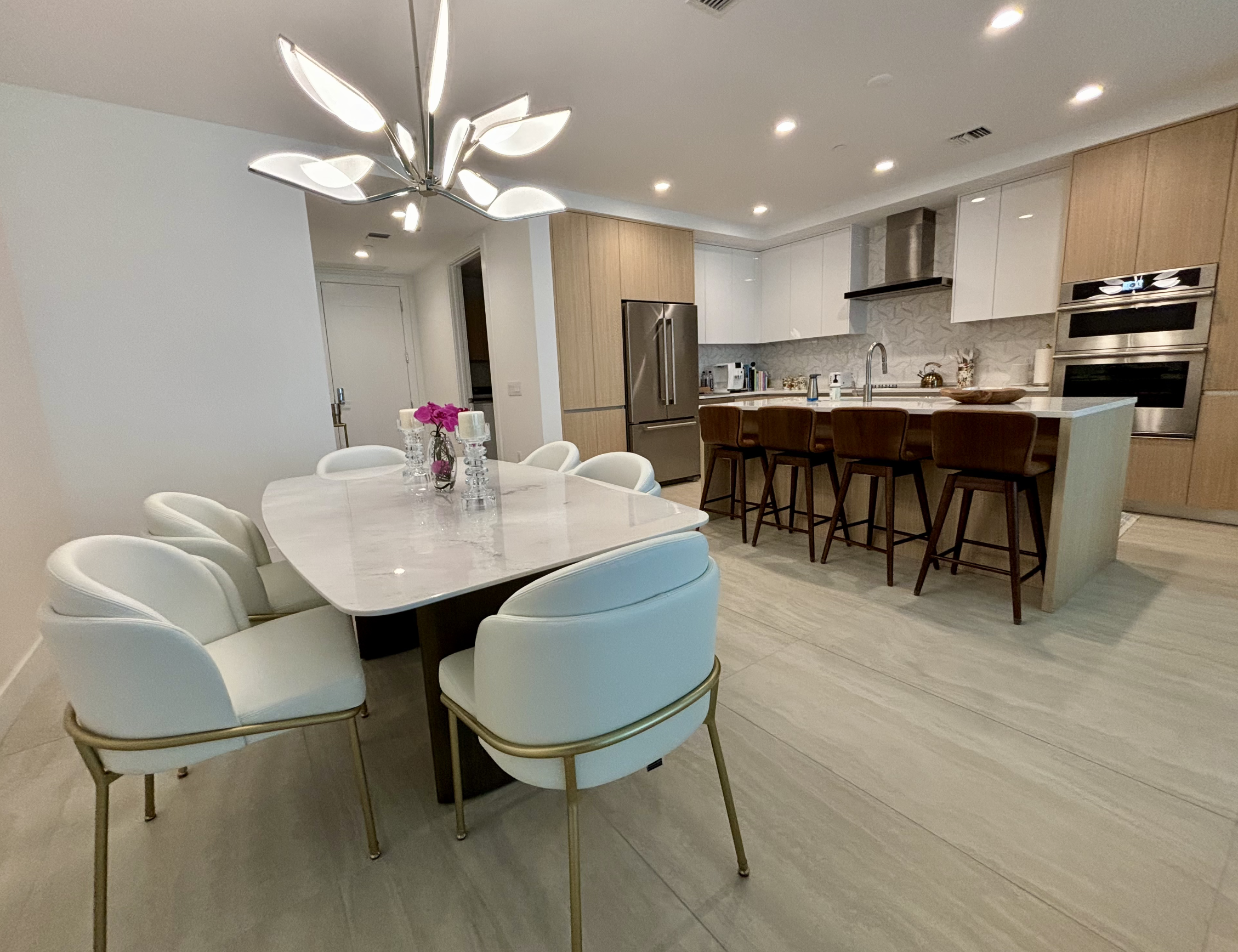 Luxury high-rise dining room at The Bayso in Sarasota featuring a Carrara marble table with a brass frame, white vegan leather chairs, and a Circa LED leaf chandelier.