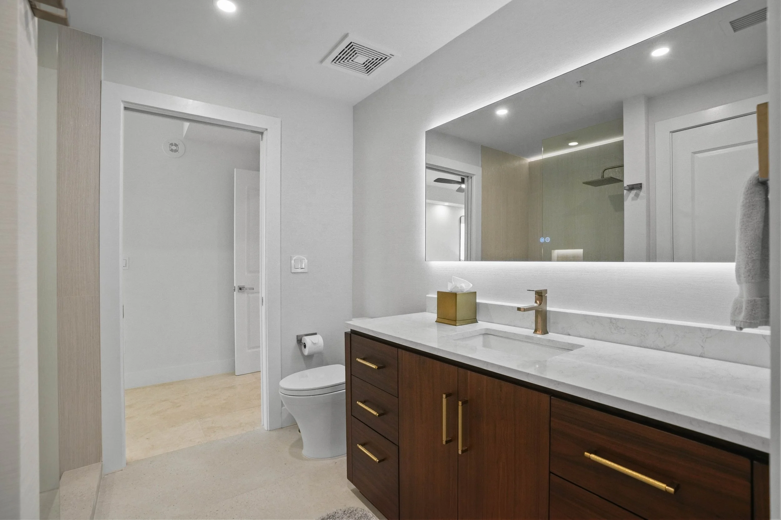 Luxury Jack and Jill bathroom remodel with walnut vanity, quartz countertop, champagne brass fixtures, Porcelanosa Japan textured tile shower, LED cove lighting, and pocket doors connecting two guest bedrooms in a Hillsboro Beach condominium.