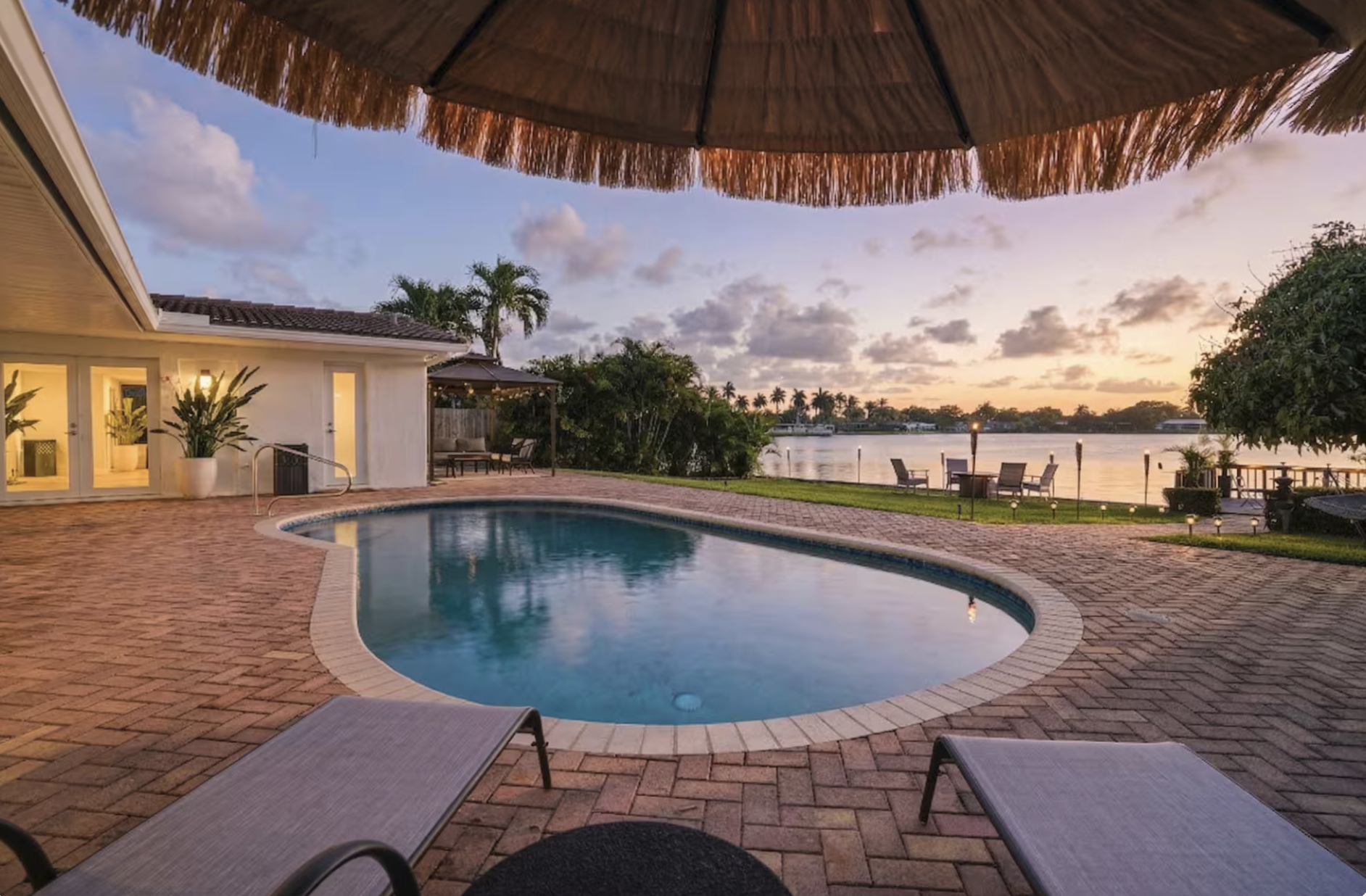 Waterfront pool patio at sunset in Miami vacation rental with lounge chairs, straw umbrella, tropical greenery, and relaxed Tulum-inspired outdoor styling.