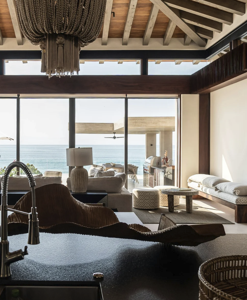 A high-end contemporary Bahamas living room featuring an architectural exposed wood ceiling, a dramatic oversized statement tassel chandelier, and towering glass doors opening to a private oceanfront patio view.