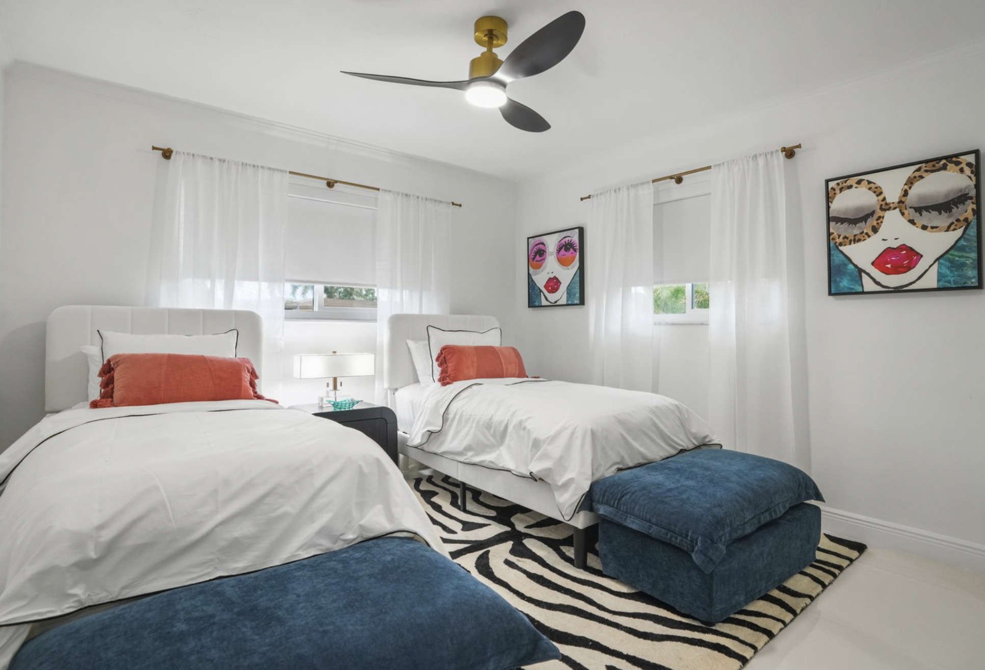 Angled view of twin guest bedroom in Miami vacation rental with upholstered beds, ottomans, graphic rug, blackout shades, and playful coastal styling.