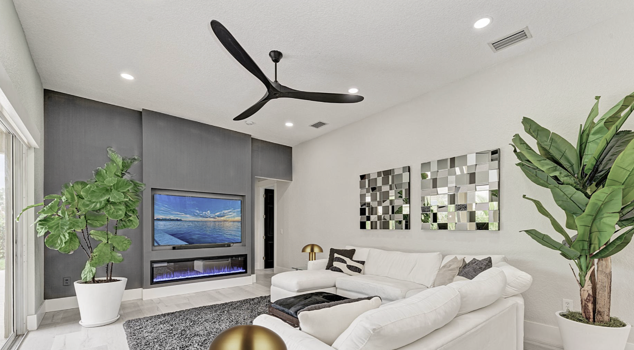 Luxury media room design in Lakewood Ranch by Exclusively to Design featuring a shimmering gunmetal chevron fireplace wall, built-in TV layout, custom haircalf ottoman, and an aeronautic ceiling fan.