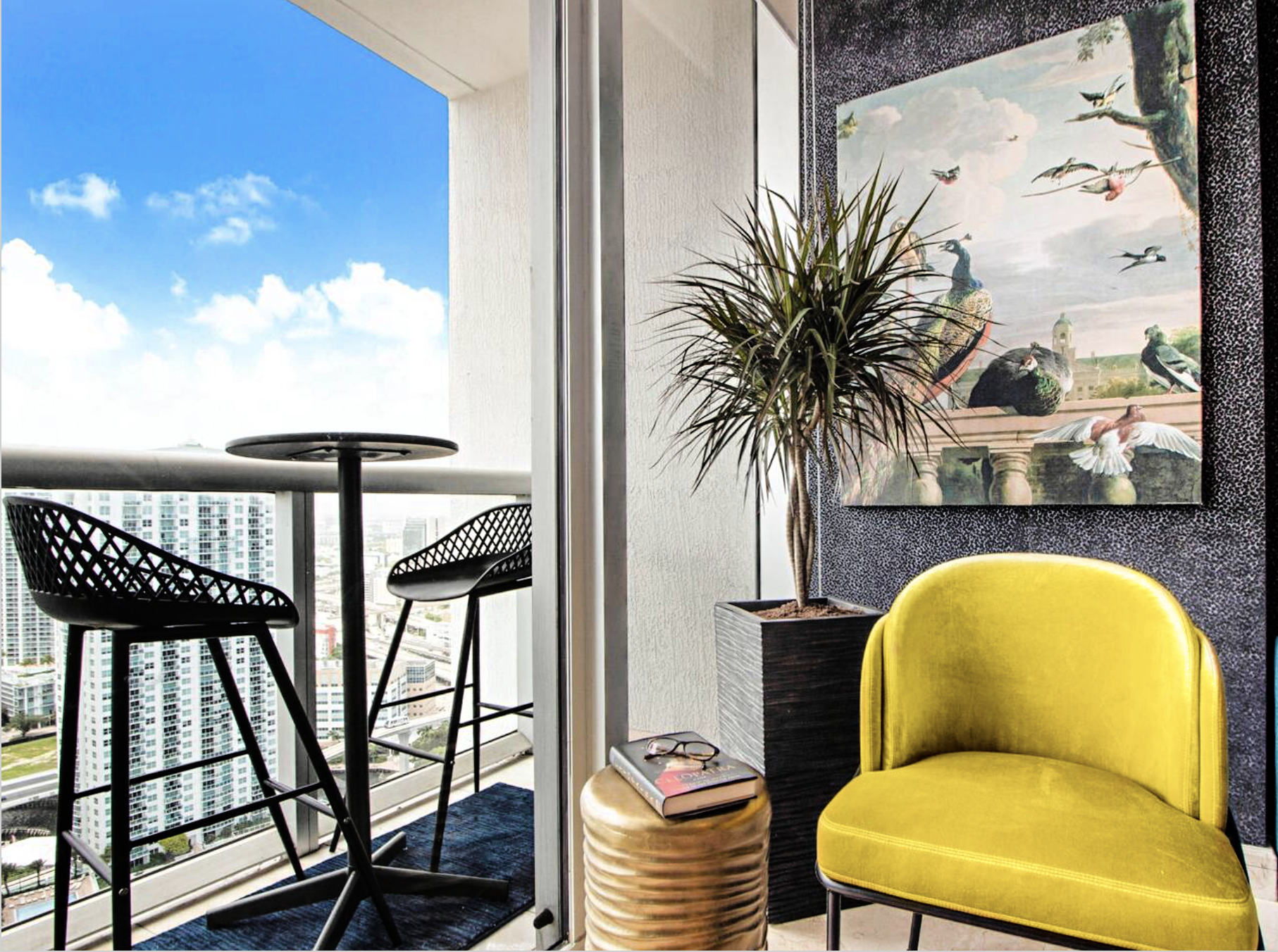 Modern balcony with two black chairs, a round table, yellow chair, potted plant, and decorative painting, offering a stunning cityscape view — by Exclusively To Design.
