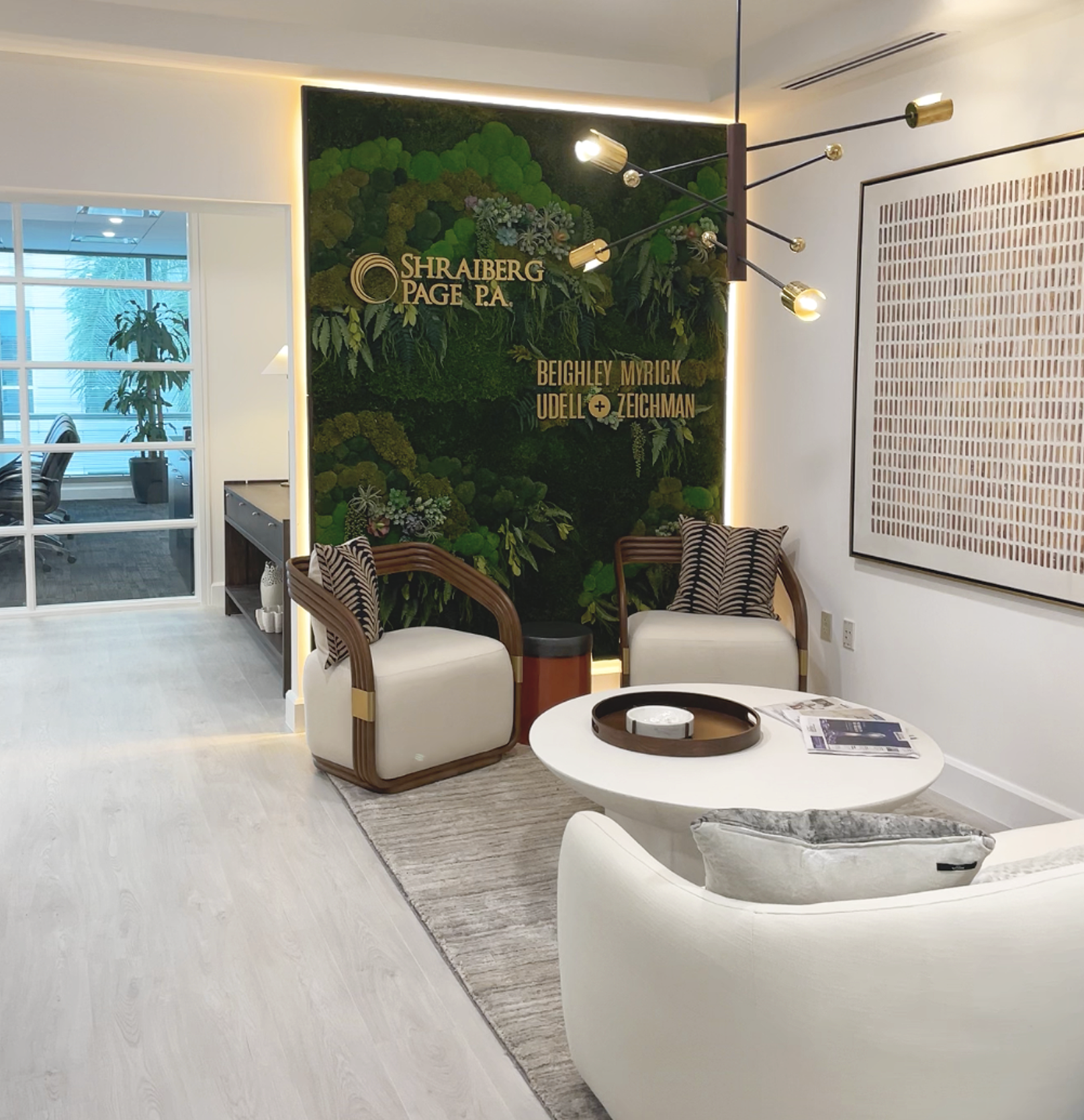 Office reception area with a green wall featuring the text 'Shraiberg Page P.A.' and the names Beighley Myrick Udel + Zeichman, modern lighting, white chairs, a round coffee table with magazines, and a framed abstract artwork on the wall.