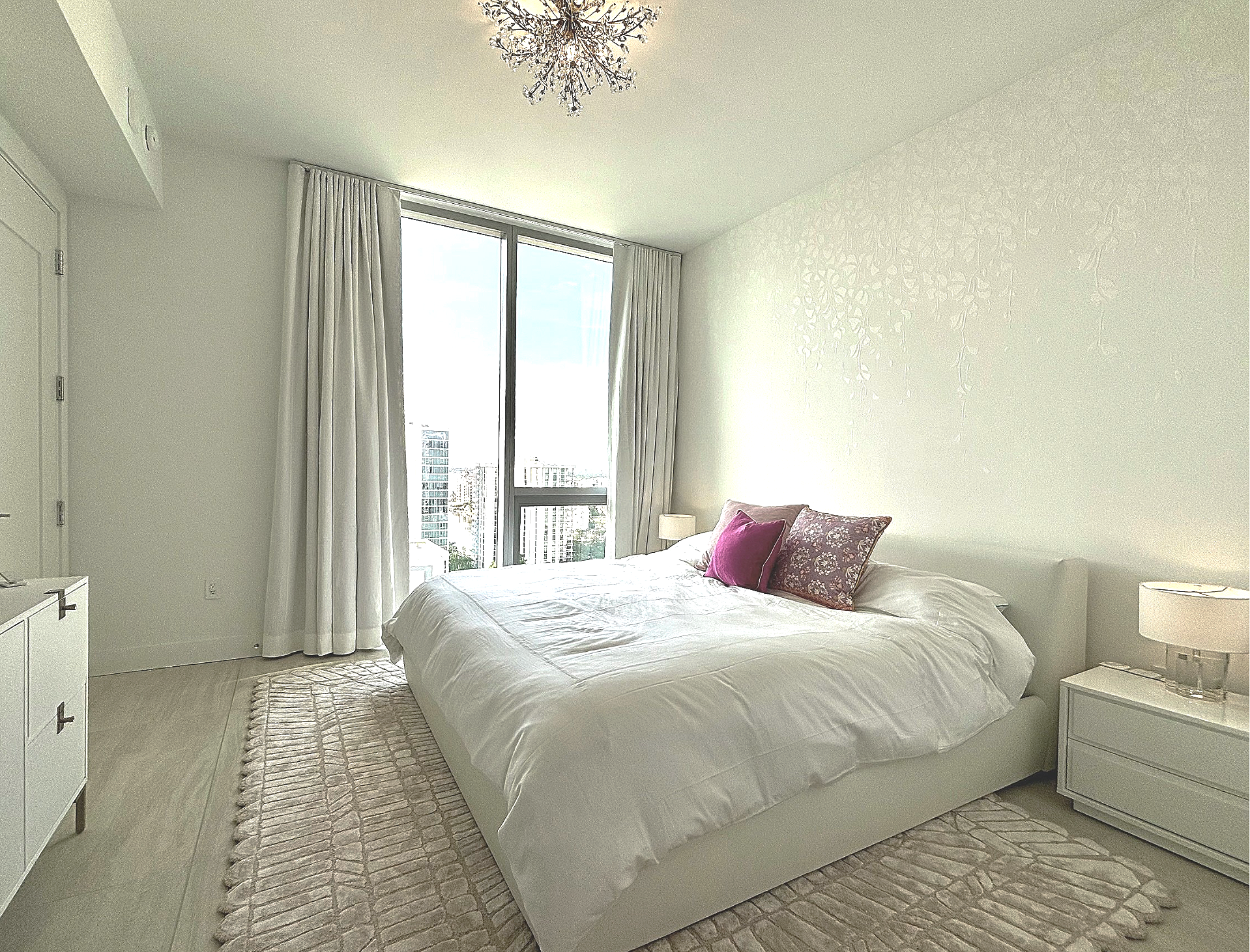 Monochromatic boho-contemporary primary bedroom design at The Baso in Sarasota by Exclusively to Design featuring a white leather bed, molded concrete falling leaves wallcovering, and crystal lighting.