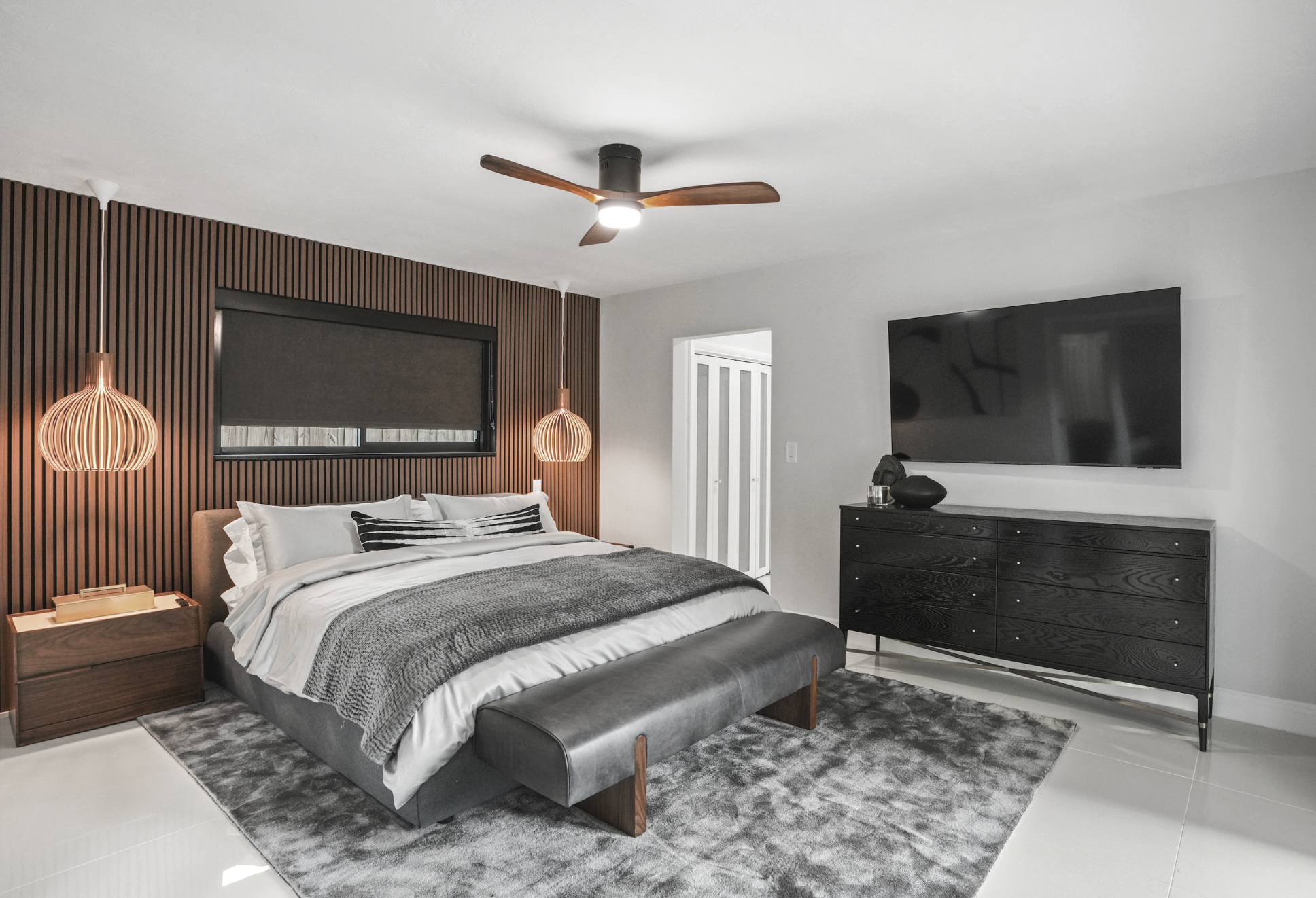 Primary suite in Miami vacation rental with walnut slat feature wall, sculptural wood pendants, blackout shade, leather bench, and masculine Tulum-inspired design.