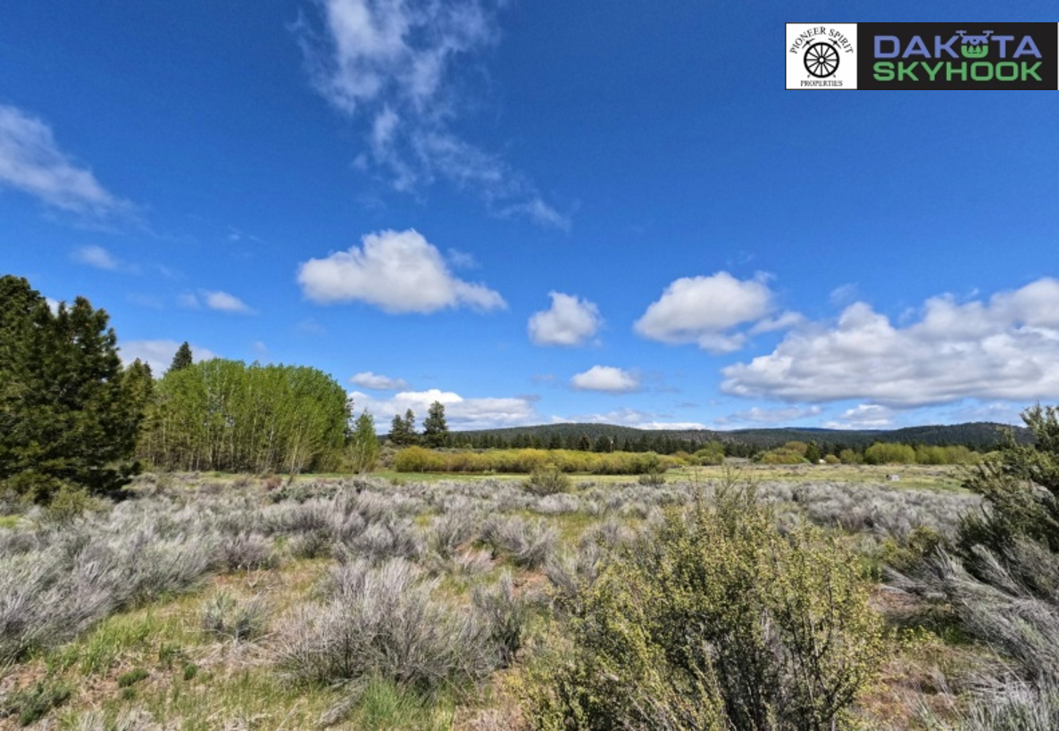 A scenic landscape with green trees, sagebrush, and open fields under a blue sky with scattered clouds.