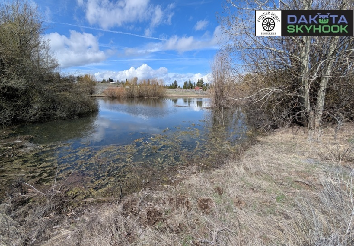 A river reflecting a partly cloudy sky, with leafless trees and dry grass along the riverbank in a rural setting.