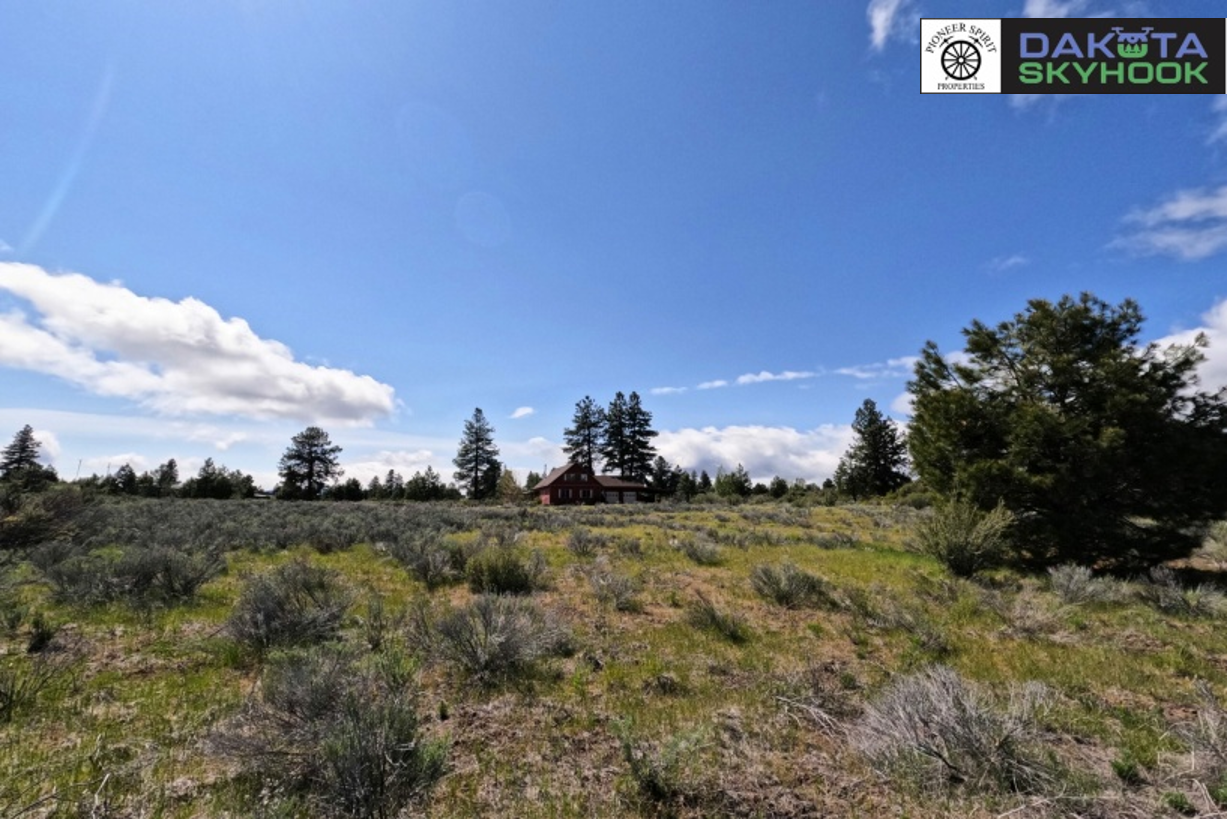 Open landscape with sparse shrubs, a few trees, a house in the distance, under a partly cloudy sky, with a logo in the top right corner reading 'Dakota Skyhook' and 'Mojer Sprt Properties'.