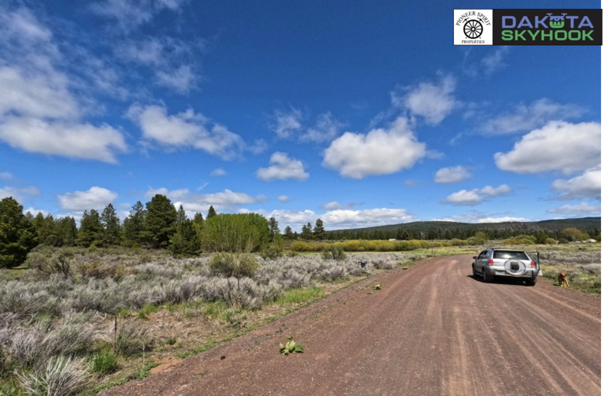 A dirt road through a rural landscape with bushes and trees, a silver car parked on the side, and a dog nearby, under a partly cloudy blue sky. Logos in the top right corner for Dakota Skyhook and Pioneer Sport Properties.