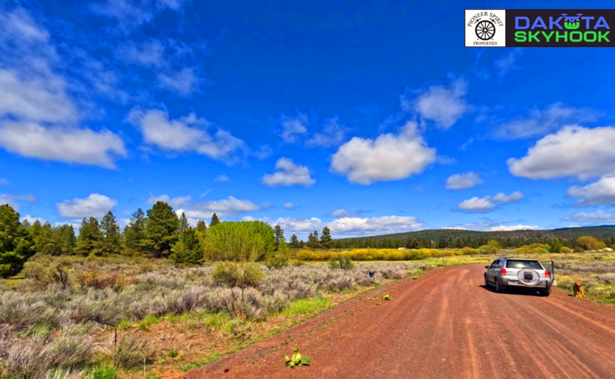 A rural dirt road with a car parked on the right side in a scenic landscape under a partly cloudy blue sky. Trees and shrubs line the horizon, with a mountain range in the distance. There is a logo in the top right corner for Dakota Skyhook.