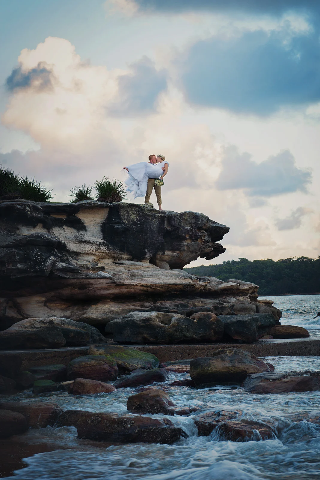 Groom holding bride in his arms on a cliff edge at Balmoral Beach, Sydney.