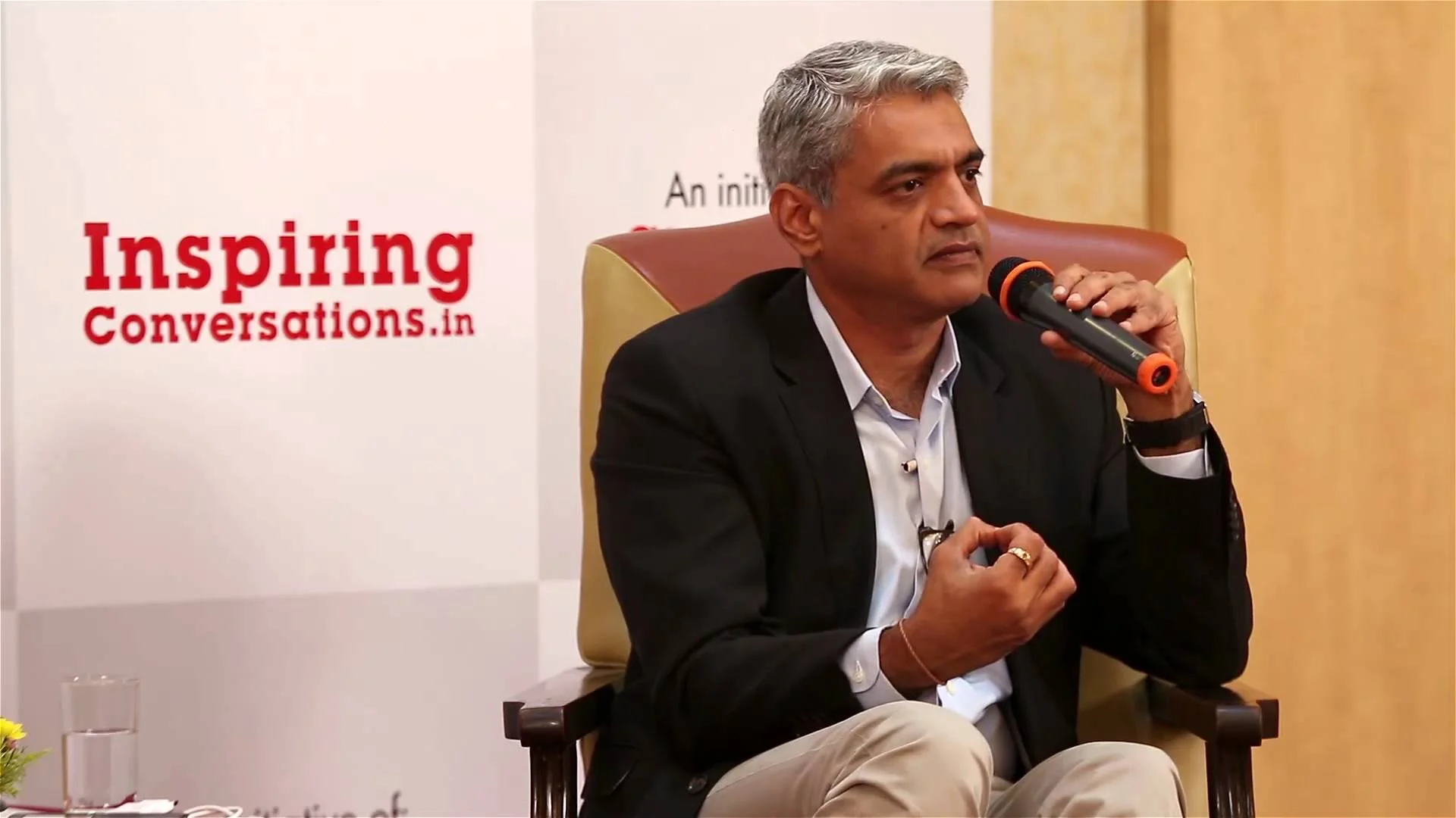Pictured above is Captain Raghu Raman - a former CEO of the Indian National Intelligence Grid and military officer, sitting in a chair while holding a microphone and acting as a guest speaker at an event. In the wall behind him there is a caption saying 'inspiring conversations.in'.