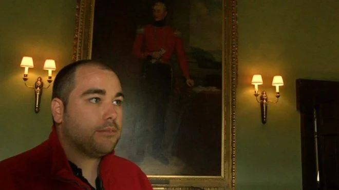 Pictured: Hayden Rustage in his red uniform, standing in front of an old painting while giving a tour of Newton House