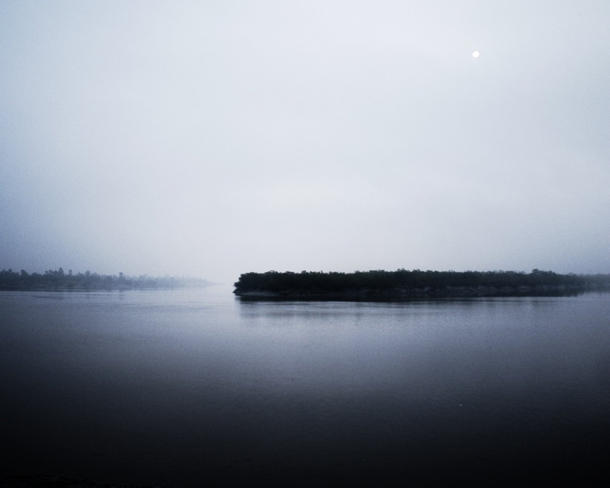 A calm body of water with an island or landmass in the distance under a cloudy sky, with the moon visible in the sky.