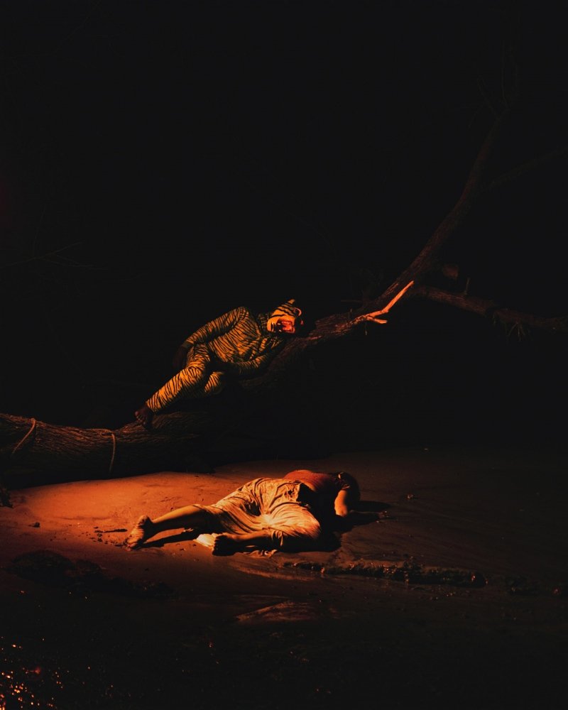 A person lying on the ground at night with a person climbing a tree nearby, illuminated by firelight or a similar warm light.