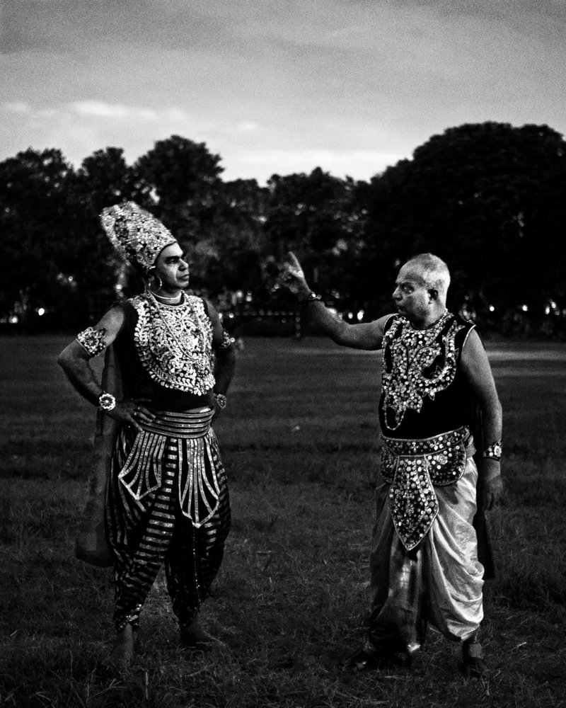 Two men dressed in traditional Indian costumes, standing outdoors on grass, with trees in the background. One man is wearing a headdress and elaborate jewelry, while the other man is raising his hand as if speaking or gesturing, both appearing to be performing or involved in a cultural event.