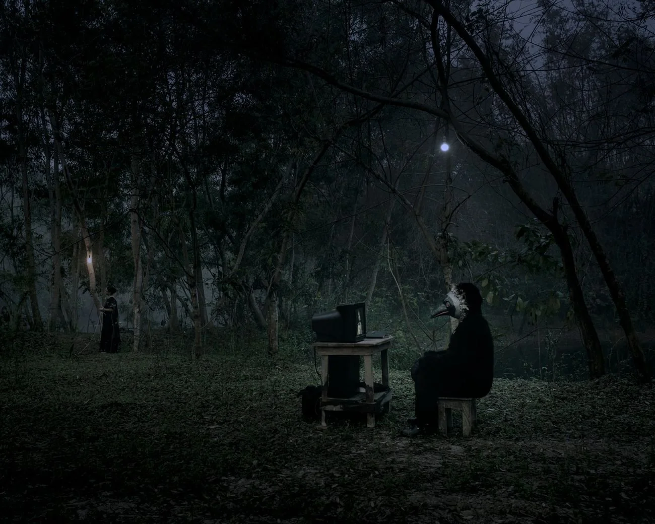 A dark forest scene at night with a person wearing a bird mask sitting in front of an old television on a small table, and another figure in the background near trees, illuminated by moonlight.