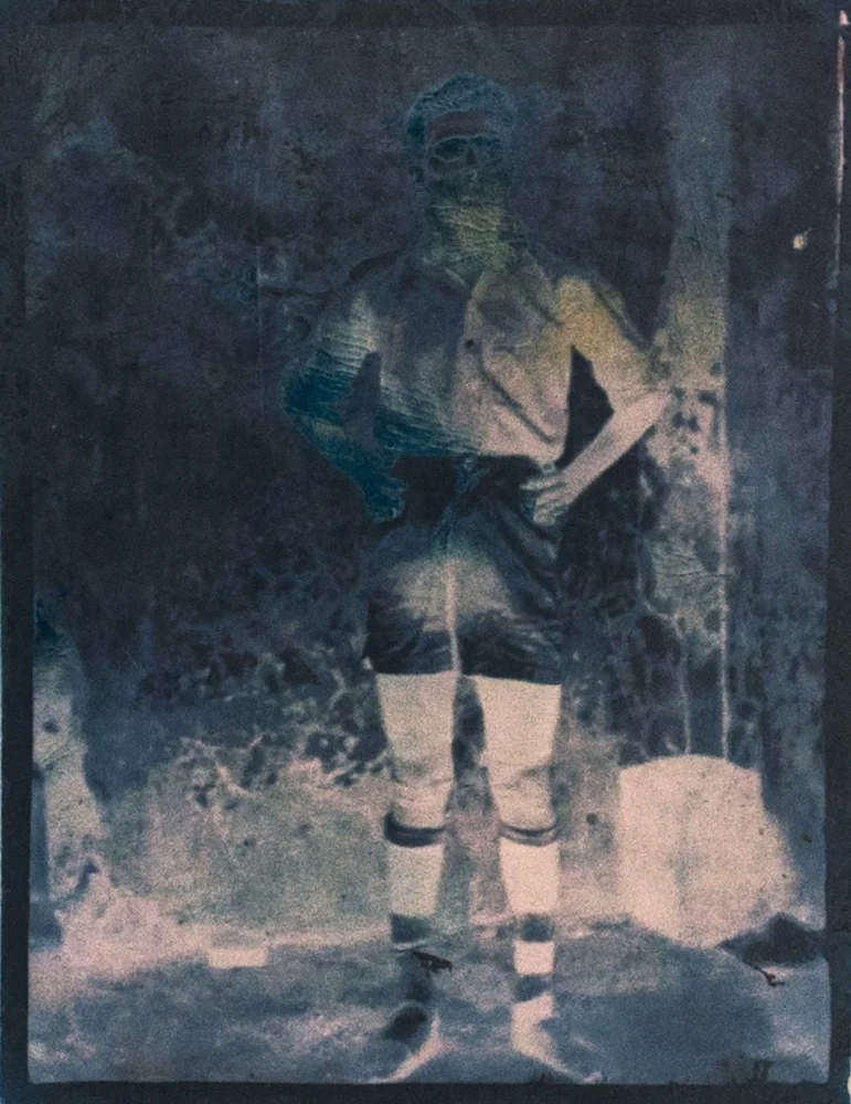 A black-and-white photograph of a man in a vintage soccer uniform with knee-high socks, standing outdoors with a hand on his hip, possibly from the early 20th century.