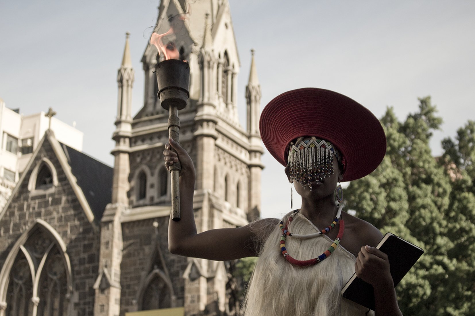 A woman dressed in traditional African attire, wearing a large red hat and beaded jewelry, holding a lit torch with a small flame in front of a historic stone church. The scene appears to be outdoors during the daytime.