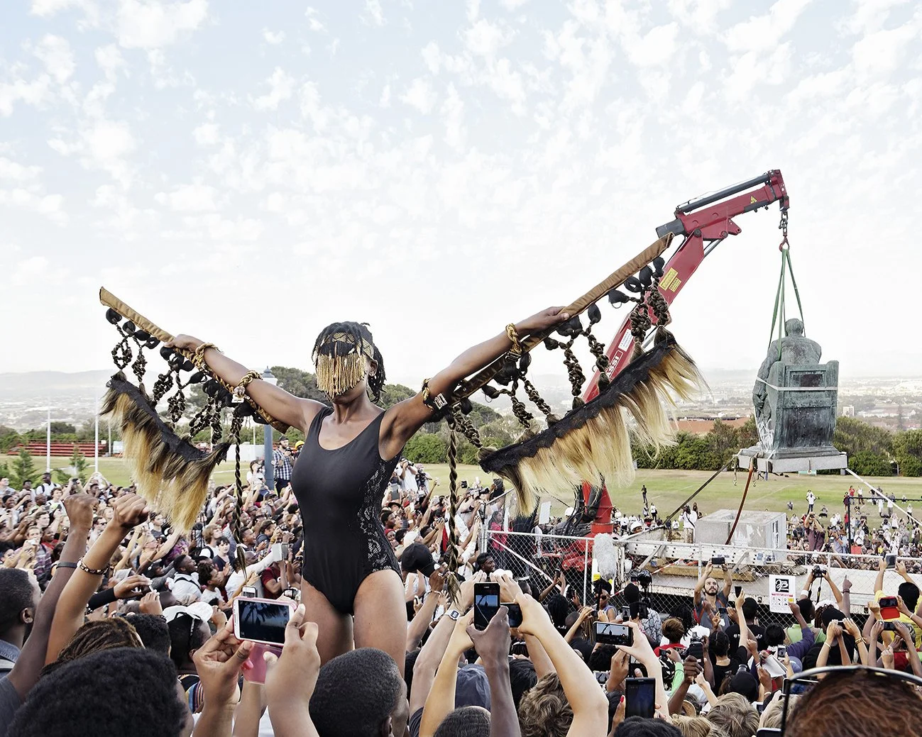 A woman with her arms outstretched, wearing a black outfit and a face covering, is present in front of a large crowd at an outdoor event. There is a large statue being lifted by a crane in the background.